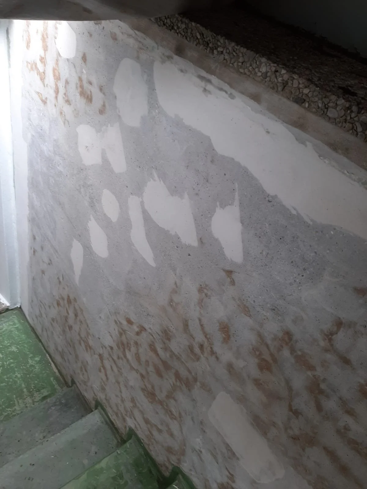 Etwa eine Wand mit frischer Putzschicht und sichtbaren Unebenheiten, neben einer Treppe und einer Gleisstrecke im Hintergrund.