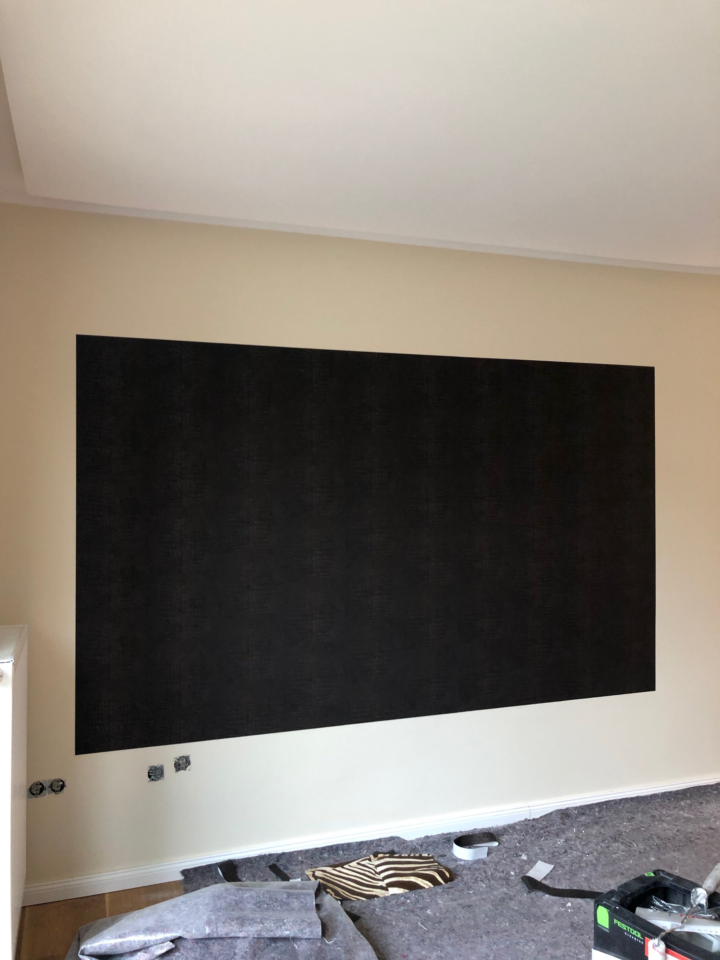 Ein Wand mit noch nicht fertiggestellt. Eine große schwarze Fläche ist auf die Wand aufgebracht. Am Boden liegen Werkzeuge und Bauteile, möglicherweise beim Streichen oder Tapezieren.
