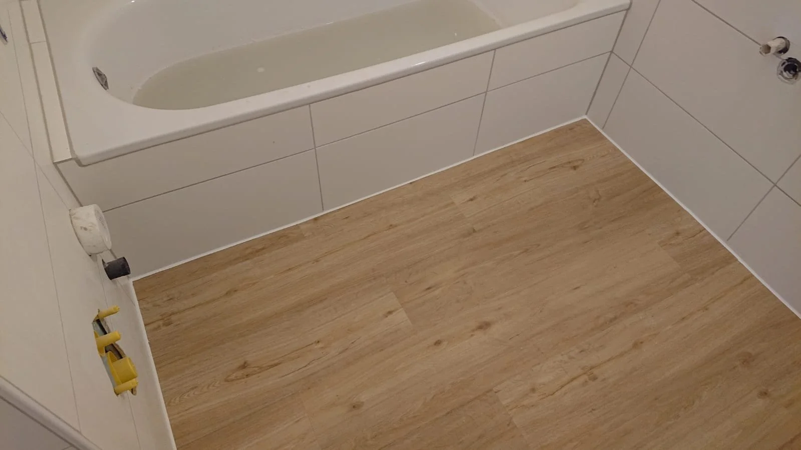 Ein Badezimmer mit Badewanne, weißen Fliesen, Holzoptik-Boden und einigen Werkzeugen auf dem Boden.