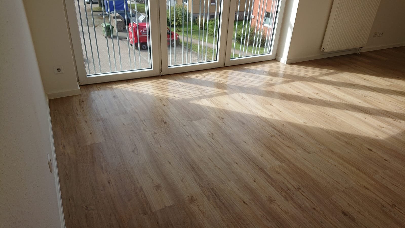 Leeres Zimmer mit Holzboden, vor einer großen Fensterfront mit einer Balkonbrüstung, durch die man einen Parkplatz und Gebäude außerhalb sehen kann.