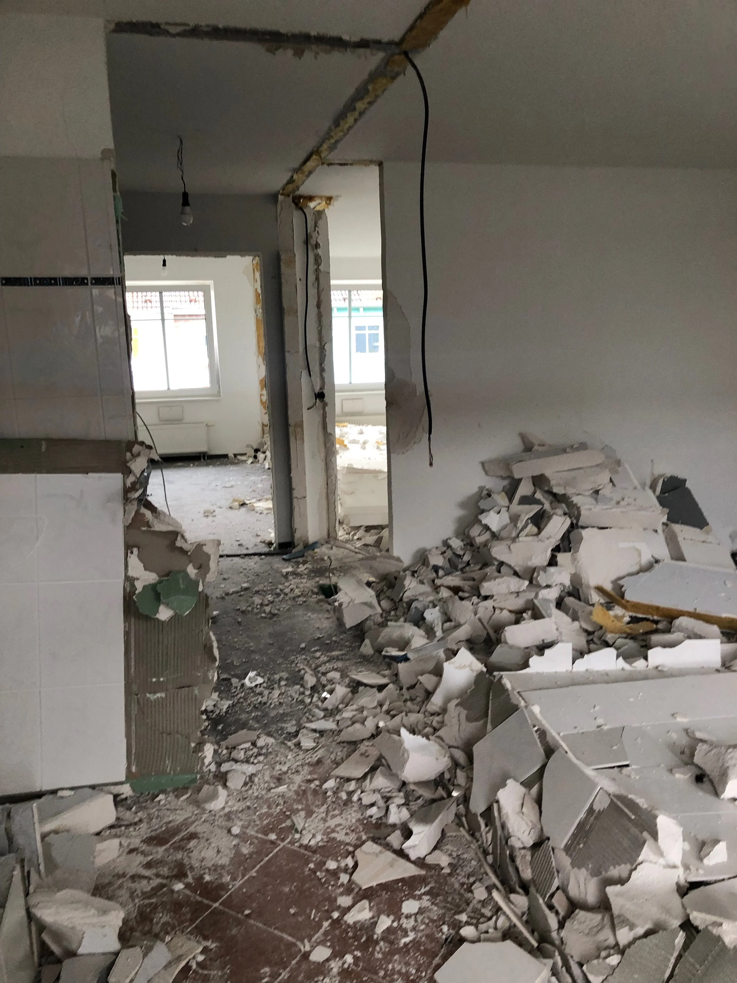 Innenansicht eines zerbombten Raums mit Trümmern und offenstehender Wand, Fenster im Hintergrund.