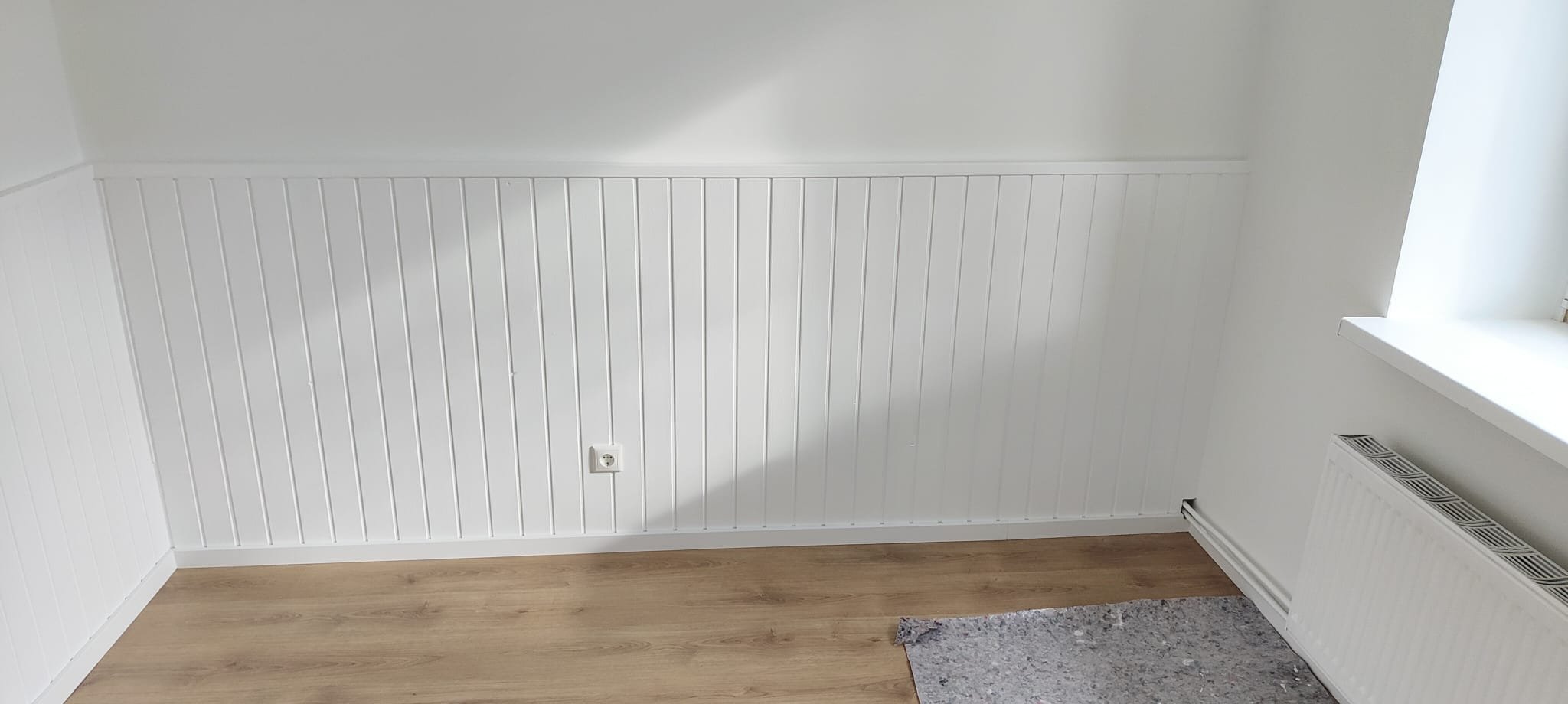 Innenraum mit weißen Wandpaneelen, einem Fenster, Heizkörper und Holzboden.