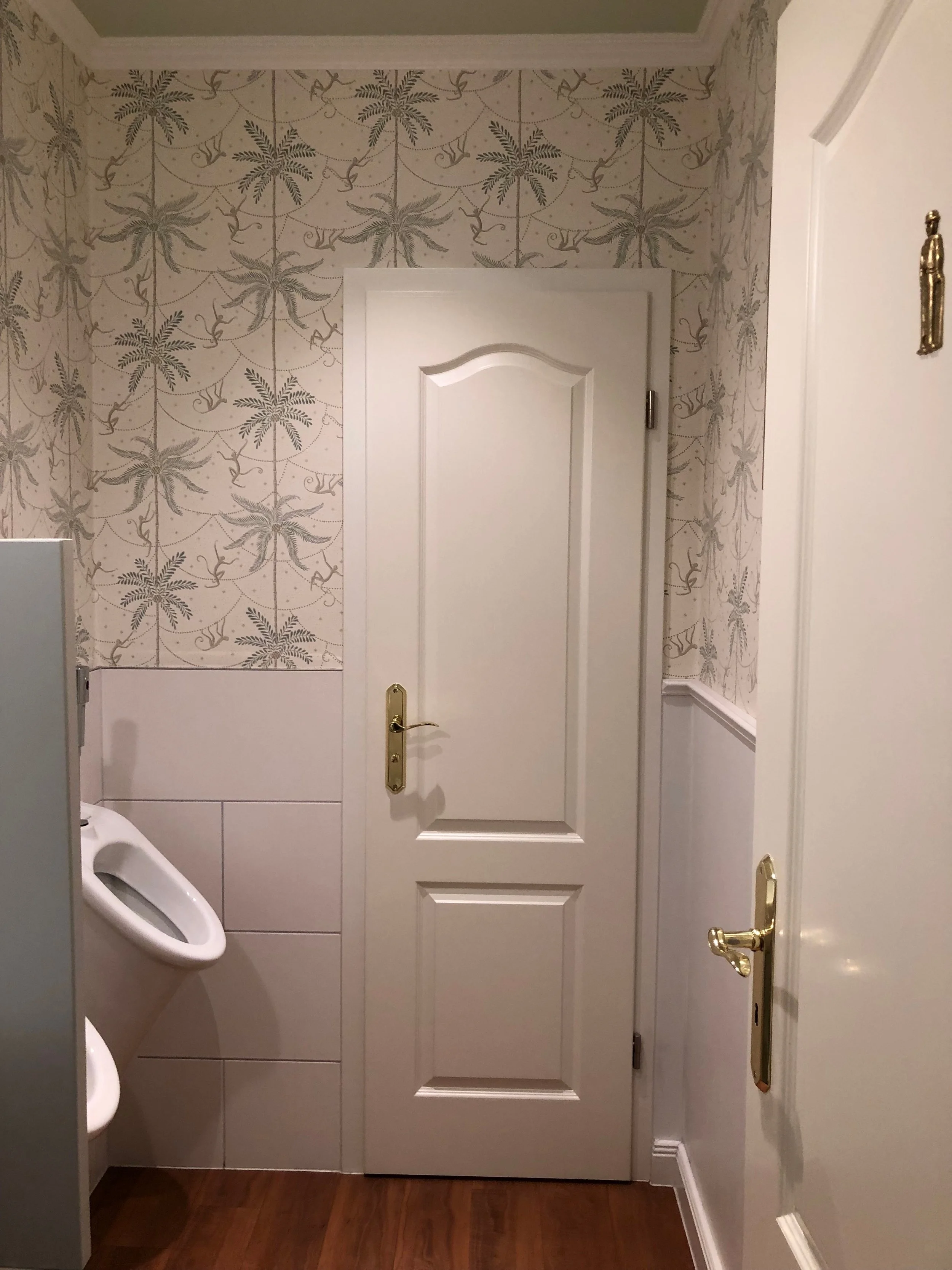 Ein Badezimmer mit Fliesen, einer weißen Tür, einer Urinal und goldfarbenen Türgriffen.