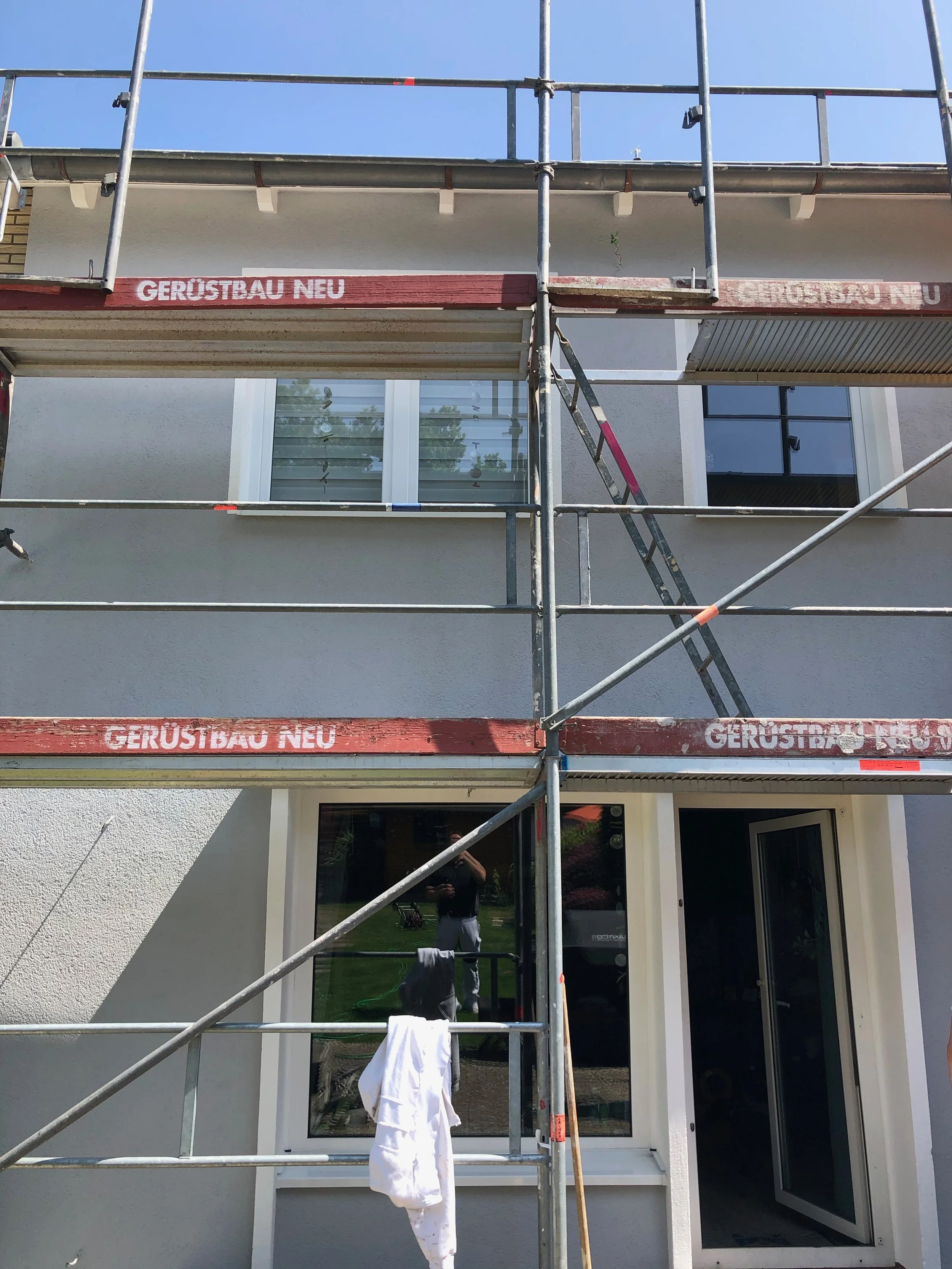 Baustelle an einem Haus mit Gerüst, das den Zugang zum Gebäude ermöglicht. Das Gerüst ist mit roten und weißen Markierungen beschriftet, die 'GERÜSTBAU NEU' sagen. Das Haus hat weiße Wände und ein Fenster sowie eine Tür, die offen steht.