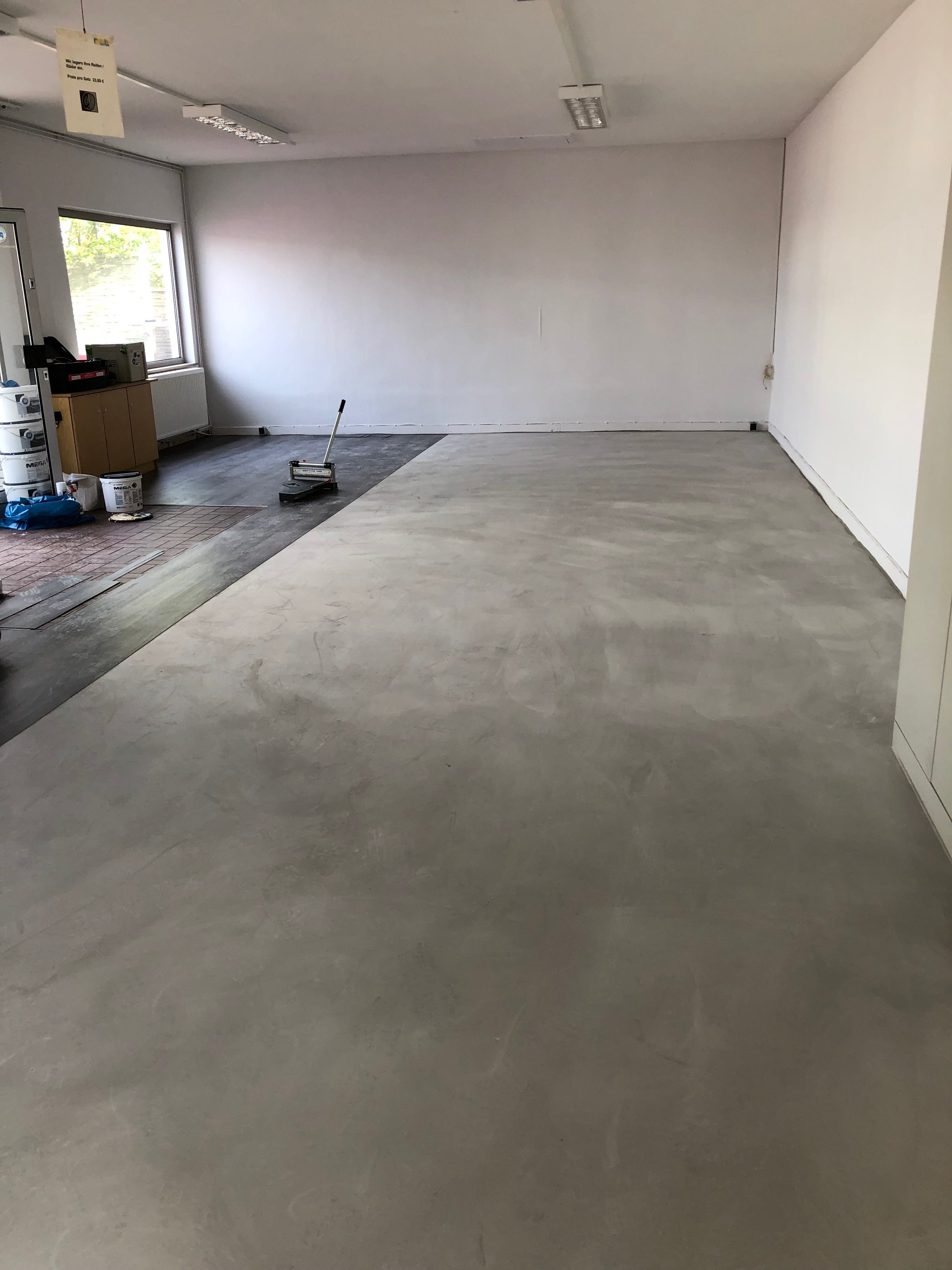 Innenraum eines Raumes mit unvollendetem Boden, teilweise ebenem, hellgrauem Betonboden, einige Baumaterialien und Werkzeuge, große Fenster an der linken Wand, weiße Wände und Decke.