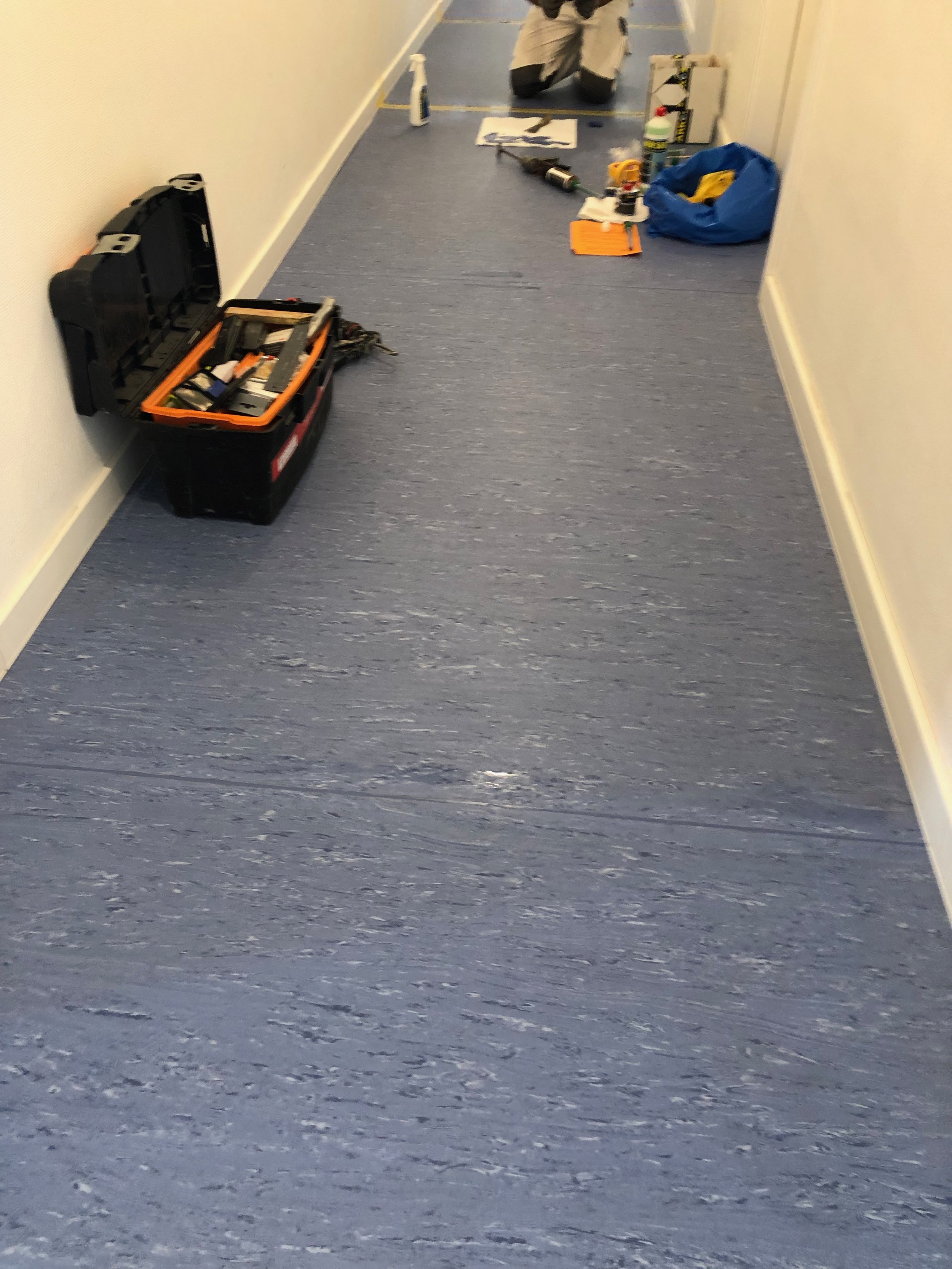 Büroflur mit Werkzeugkasten und Werkzeugen, Reinigungsmittel und einem blauen Beutel auf dem Boden, im Hintergrund eine Person mit einem Akkuschrauber
