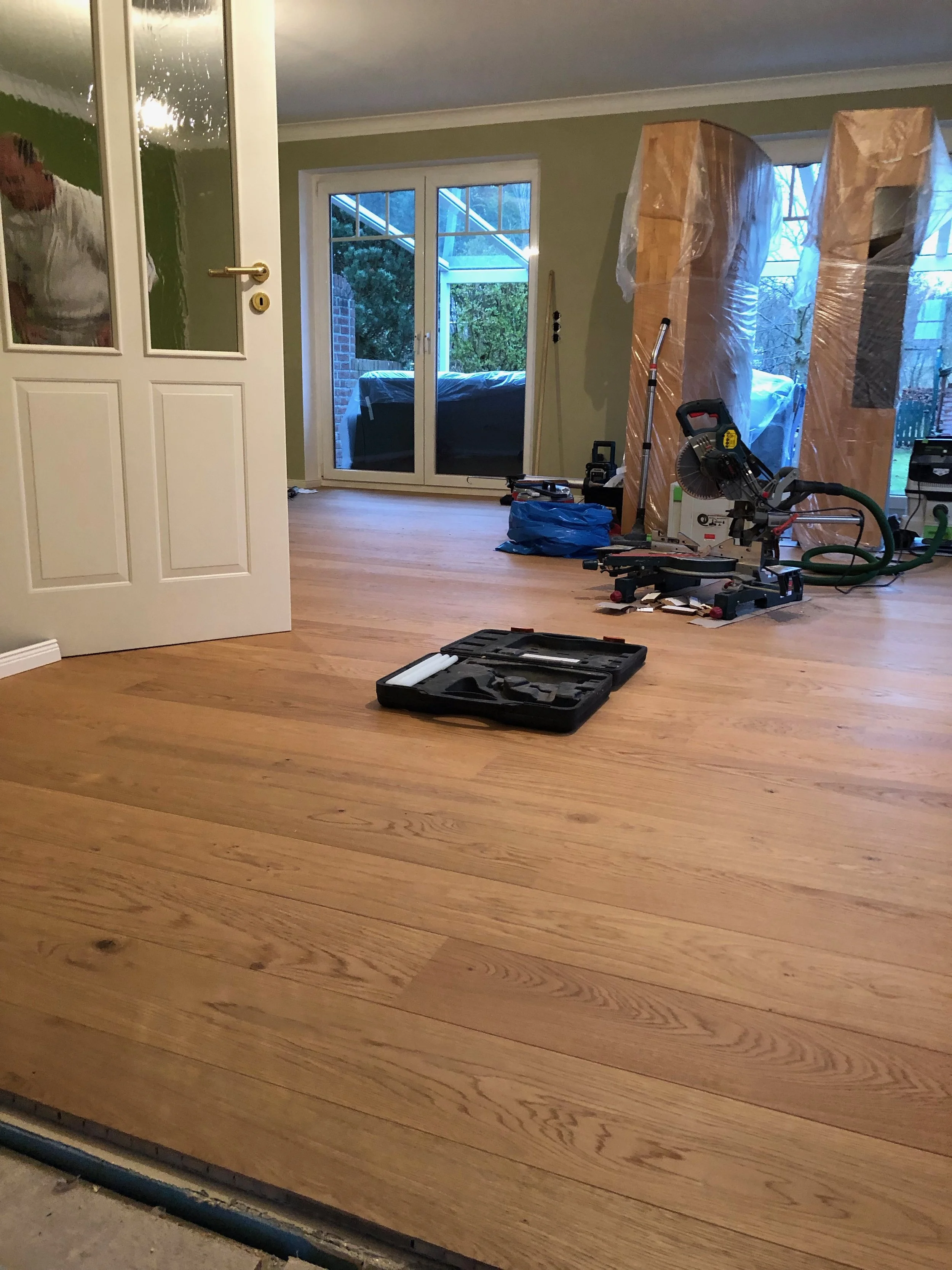 Innenaufnahme eines Wohnzimmers während einer Renovierung, mit geöffnetem Tür, Holzfußboden, Werkzeuge und Möbel vorübergehend abgedeckt.