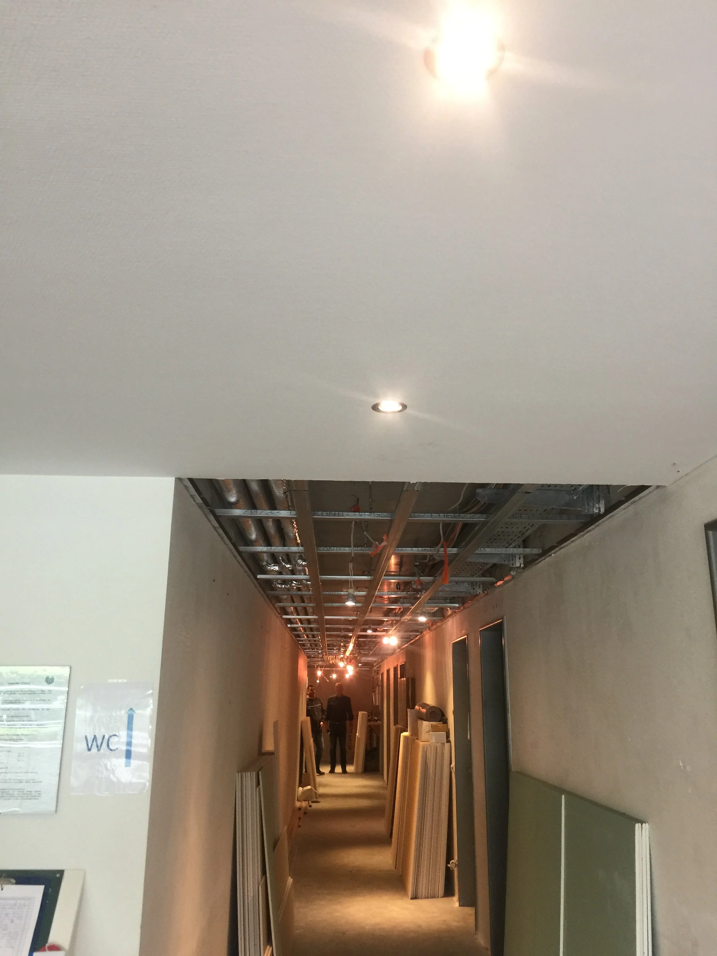 Innenansicht eines Flurs in einem Gebäude, das noch gebaut oder renoviert wird, mit sichtbaren Deckeninstallationen, Baustoffen und ein paar Personen im Hintergrund.