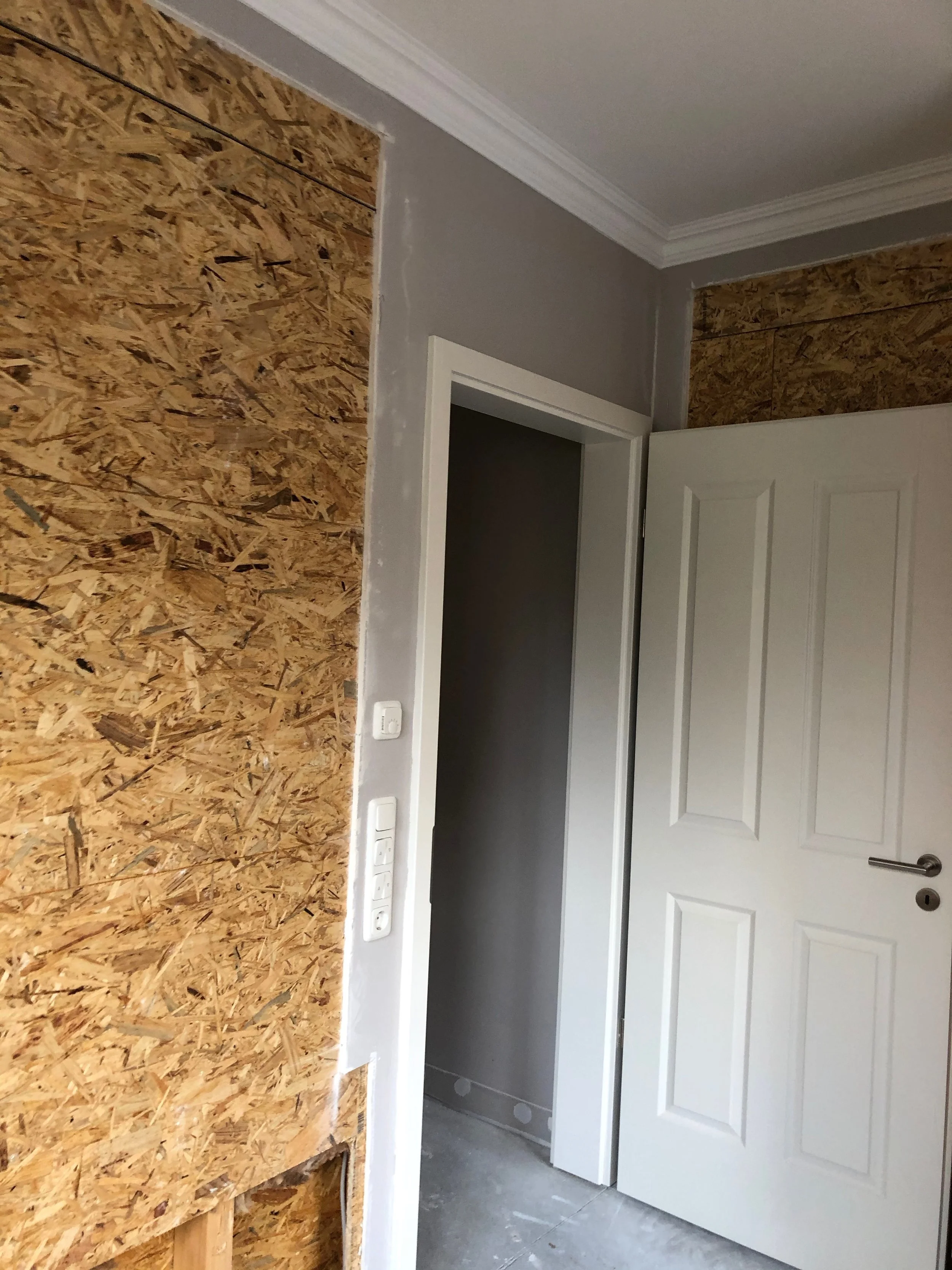 Ein Raum im Bau mit einer weißen Tür, einer Wandverkleidung aus Sperrholzplatten und einem Lichtschalter