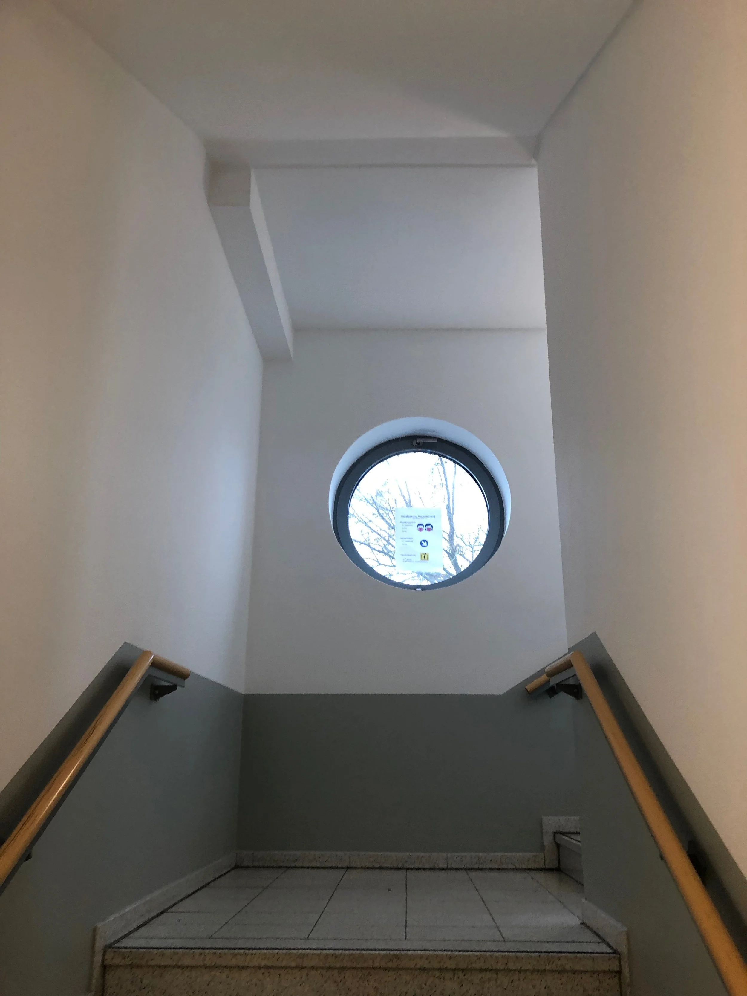 Innenaufnahme eines Treppenhauses mit grauen Wänden, Holzhandläufen und einer runden Fensteröffnung, durch die ein Baum und der Himmel zu sehen sind.