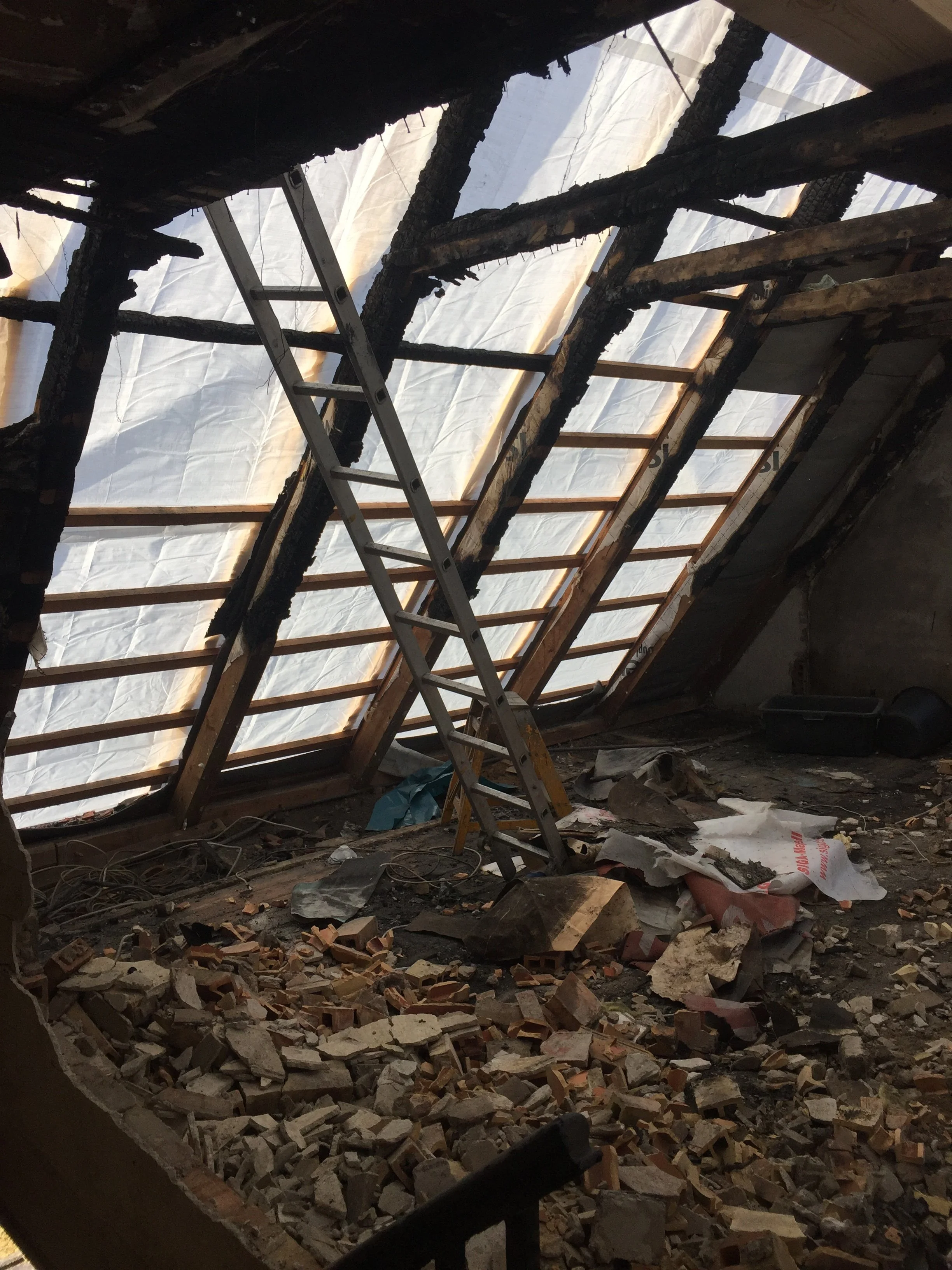 Innenansicht eines Dachbodens mit beschädigtem Dach, Schutt und Baumaterial, eine Leiter liegt beim Umklettern