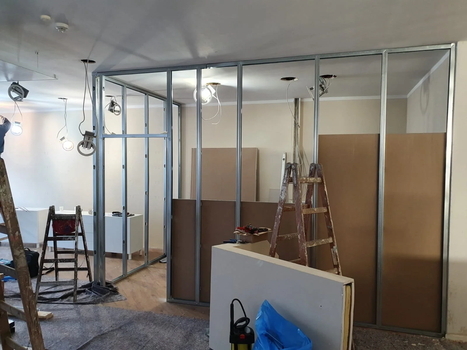 Innenaufnahme eines Raumes in Renovierung, mit Metallrahmen für Wände, Leitern und Baumaterialien, Hängende Lichtquellen und unvollständiger Decke.