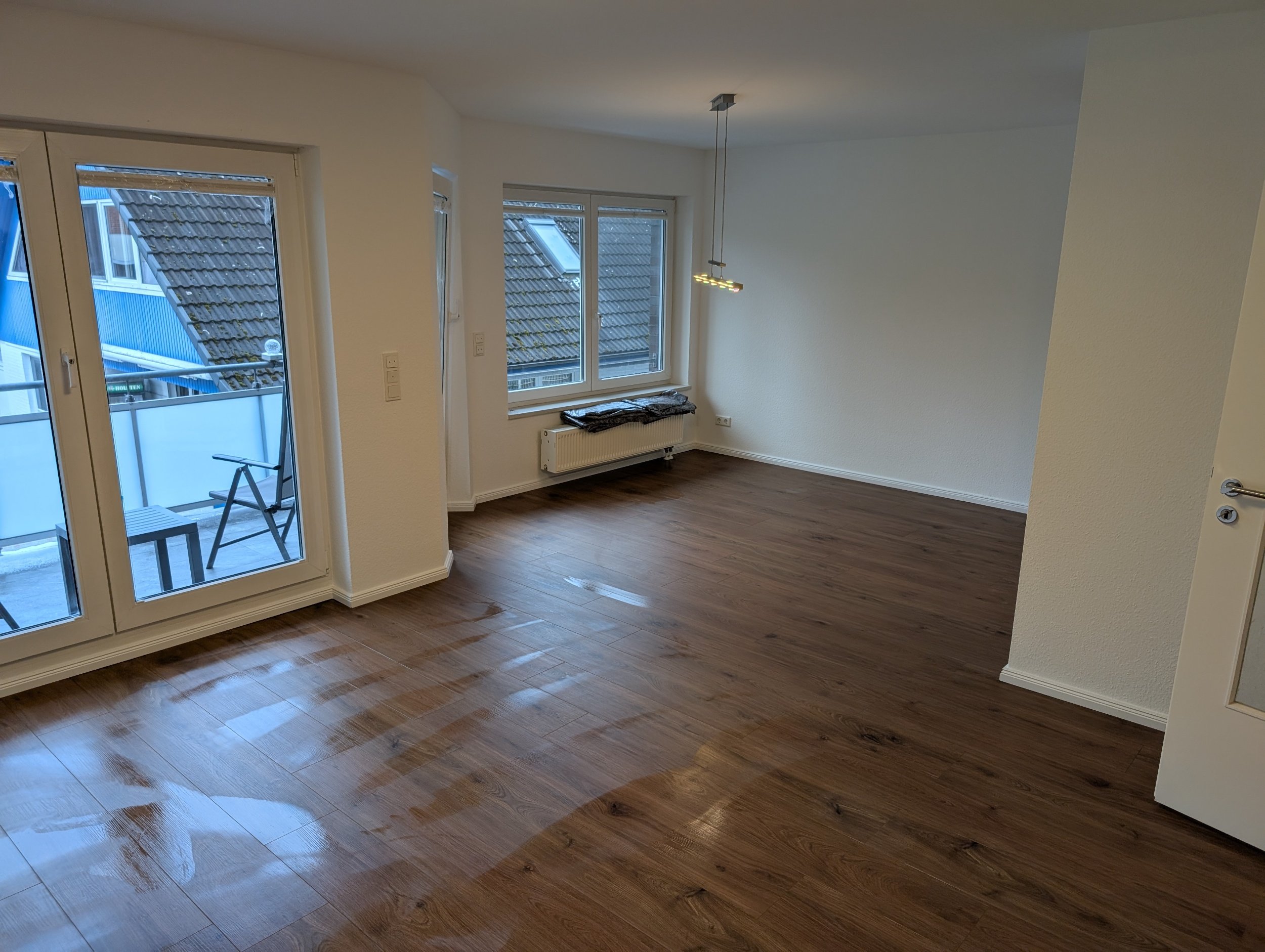 Ein leerer, moderner Wohnraum mit Holzboden, großen Fenstern und Zugang zu einem Balkon.