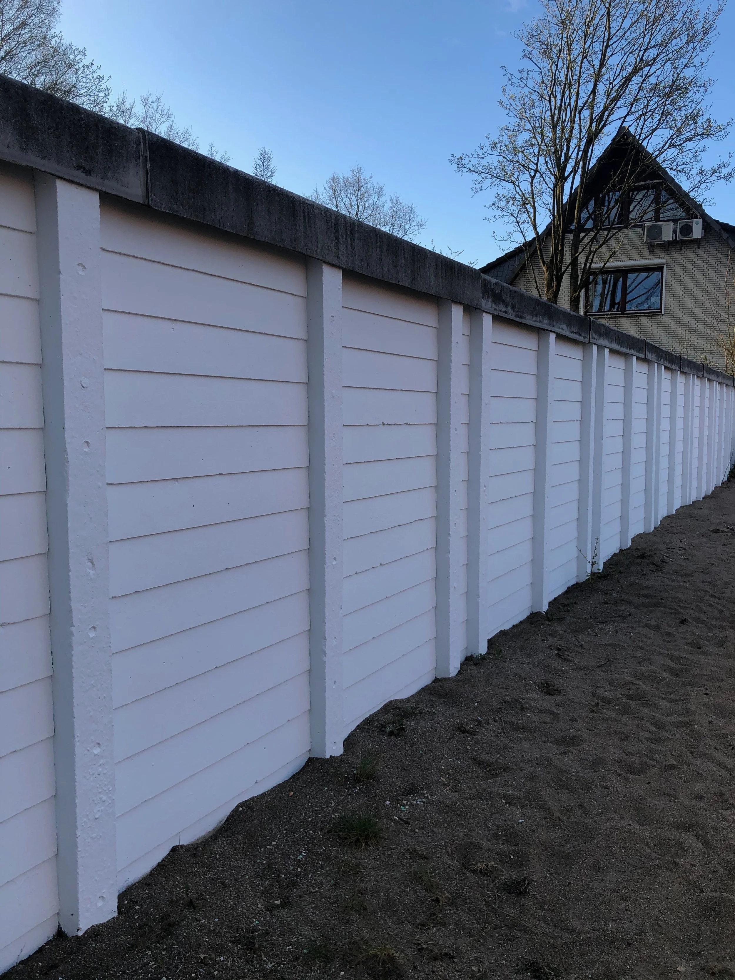 Weißer Lattenzaun auf einem dunklen Boden, im Hintergrund ein Haus mit Dach und Baum.