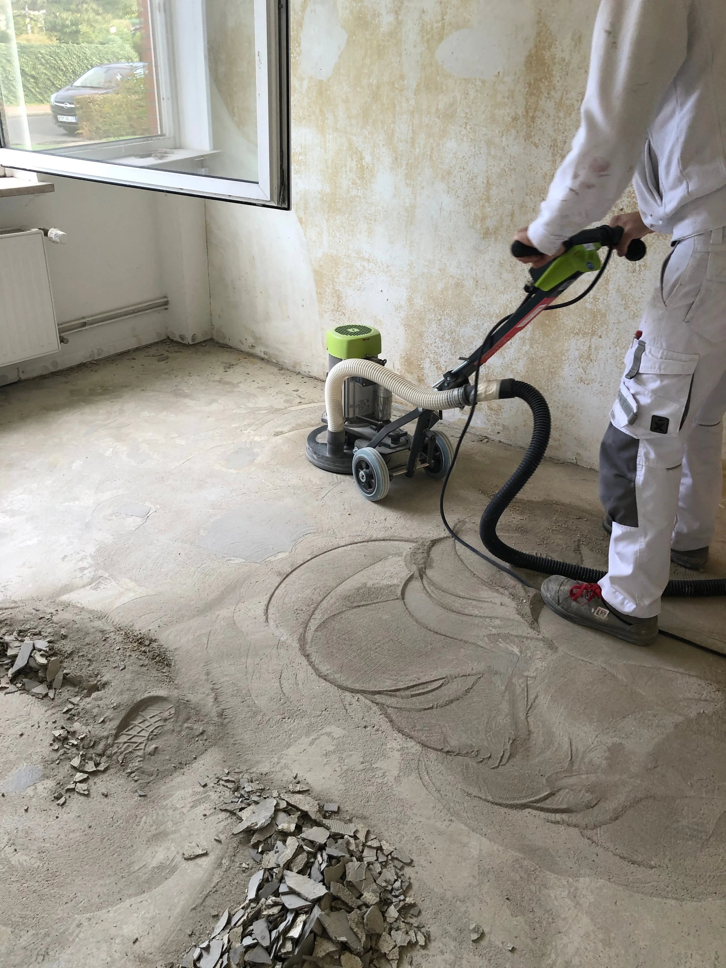 Ein Handwerker im weißen Arbeitsoutfit benutzt ein Schleifgerät auf einem alten Fußboden, um ihn zu renovieren.