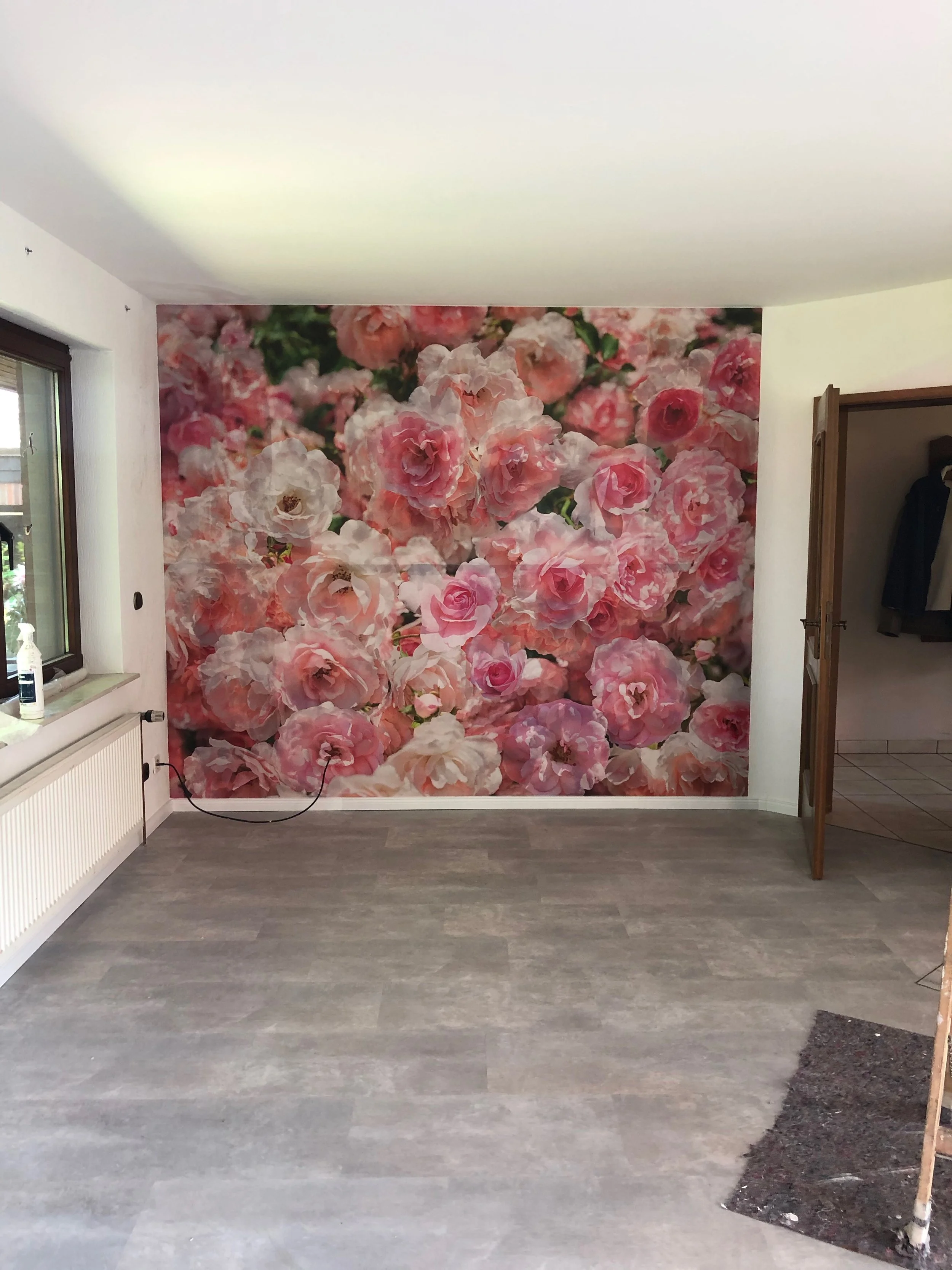 Ein Raum mit einer großen Wandtapete, die rosa und weiße Rosen zeigt. Der Raum hat einen Holzboden, eine Fensterbank mit einem Desinfektionsmittel und ein offenes Tür, die in einen Flur führt.