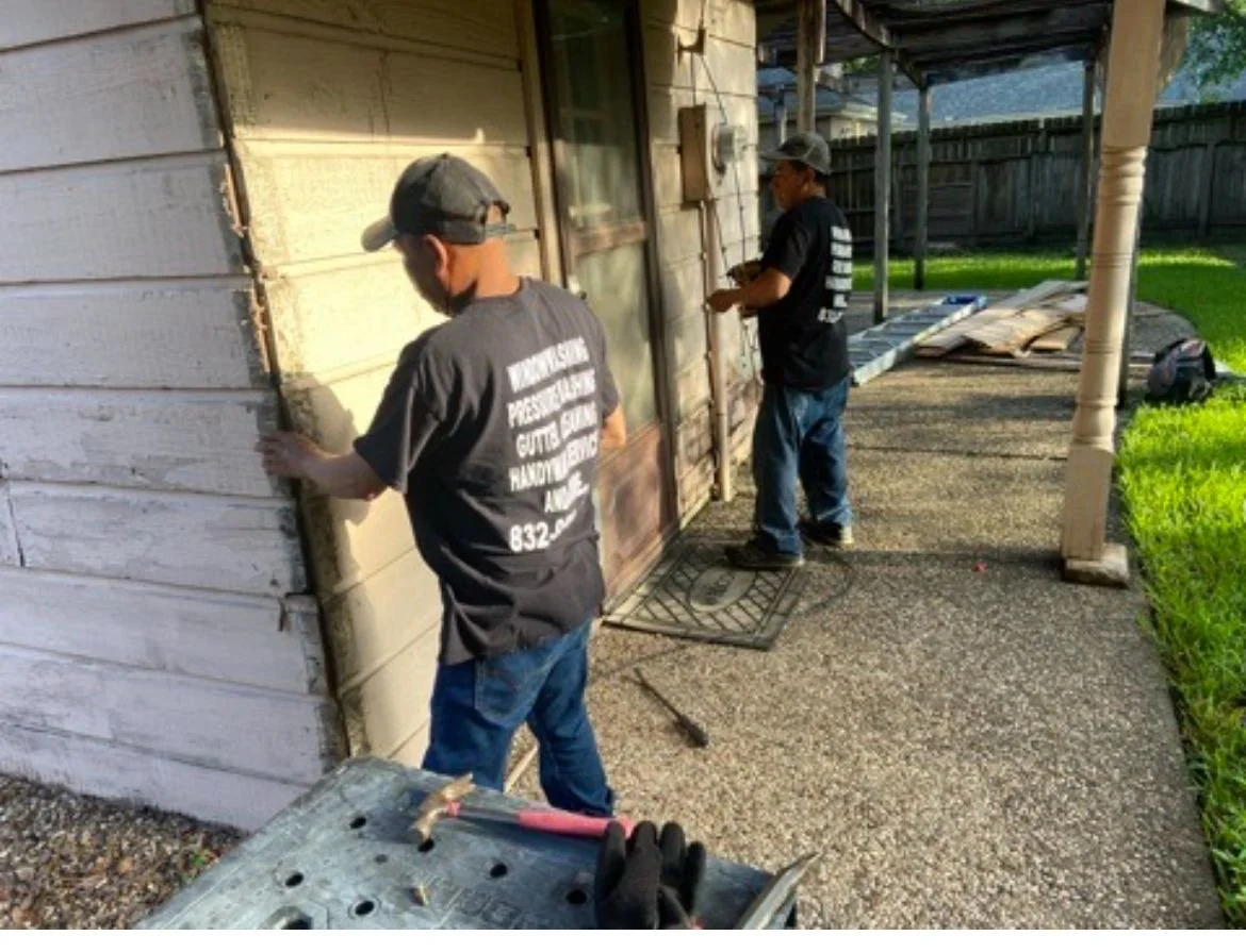 Two men performing home repairs on a wooden exterior, with tools and materials on the ground, on a sunny day.