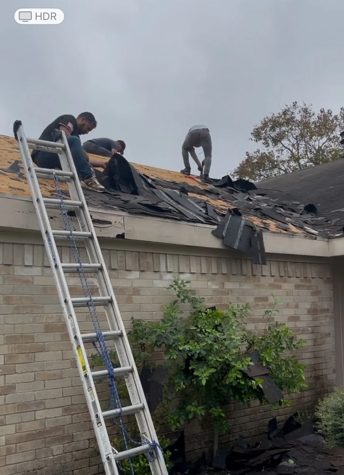 Three people working on roof repair, with a ladder propped against a brick house.