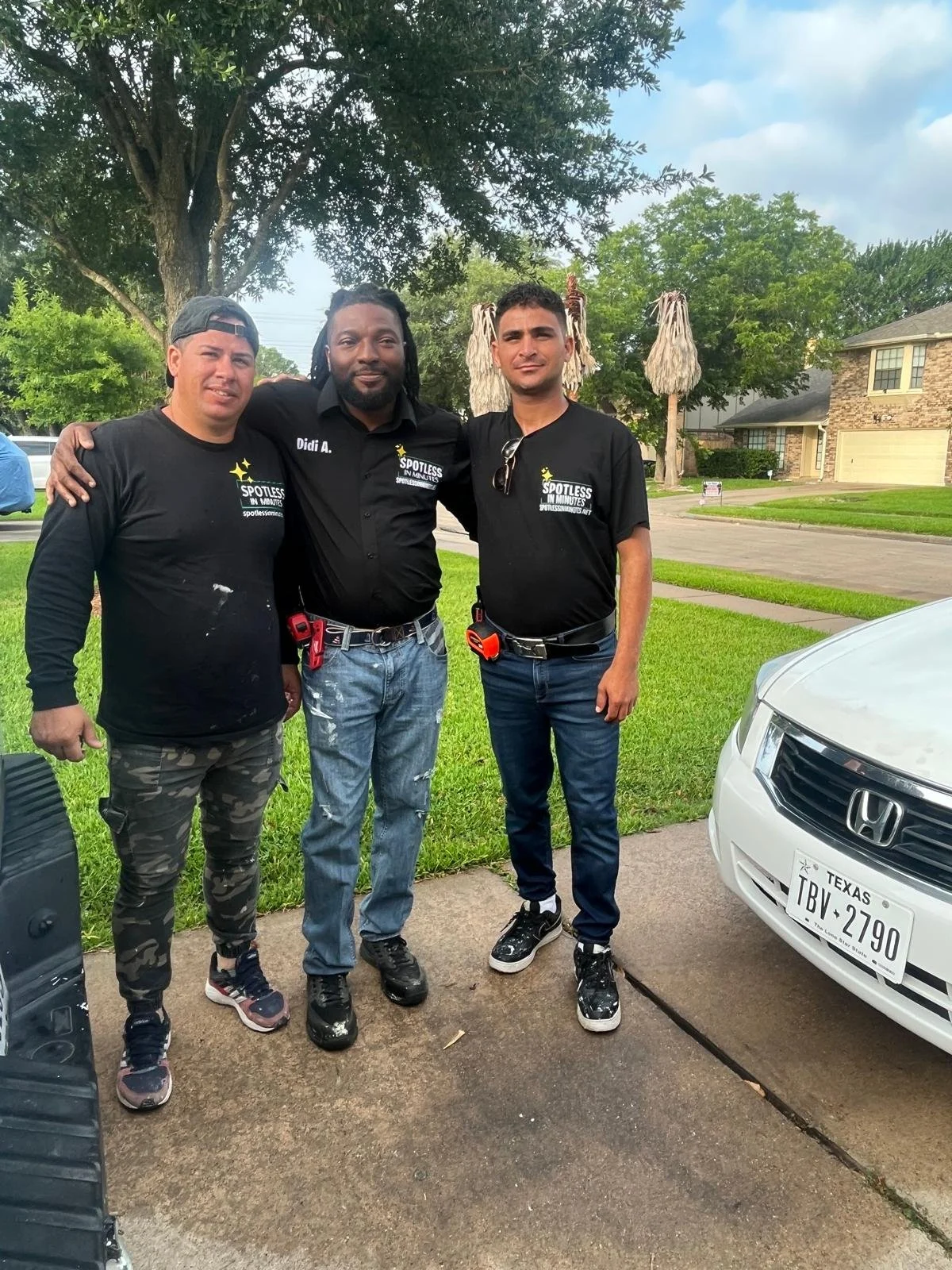 Three men wearing matching "Spotless Windows" shirts pose for a photo next to a car with a Texas license plate in a suburban neighborhood.