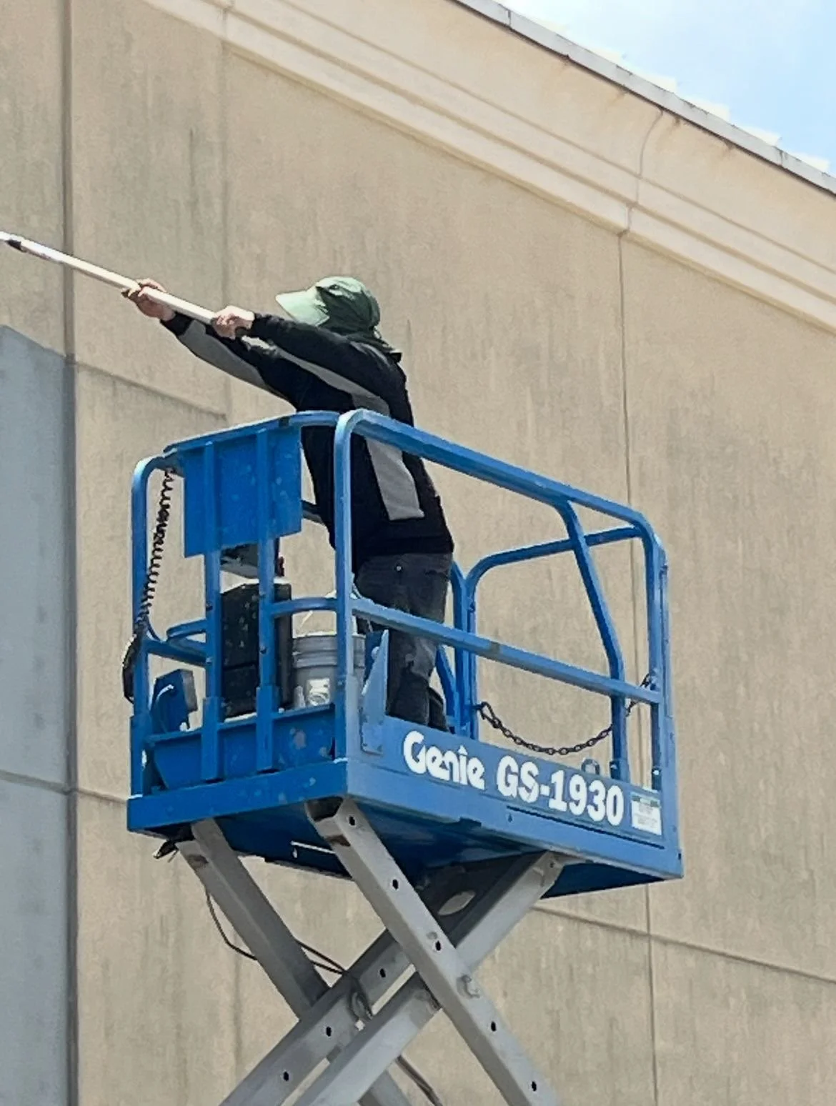 Person painting a building wall from a scissor lift, wearing a hat, using a long roller brush.