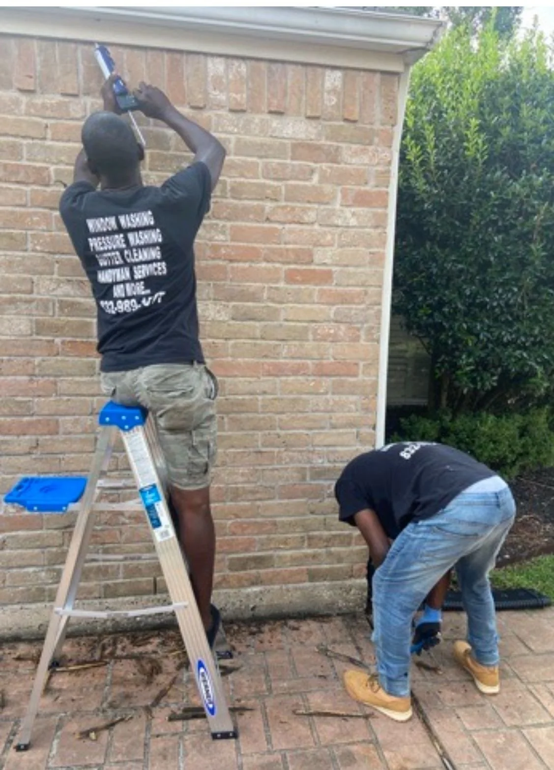 Two people working on a brick wall, one on a ladder using a caulking gun, the other fixing something near the ground, both wearing work clothes.