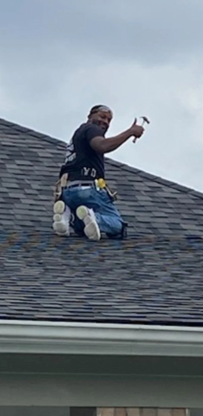 Person kneeling on a roof with a tool belt, holding a hammer, working on shingles.