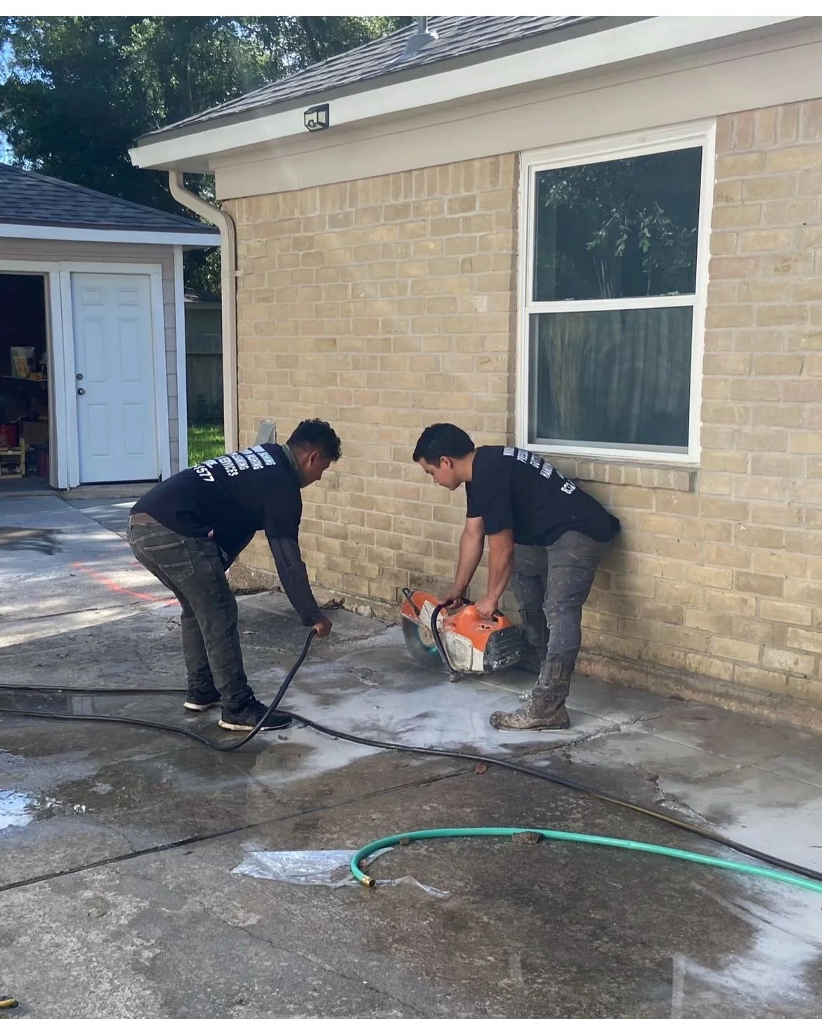 Two workers using a concrete saw on a driveway next to a brick house.
