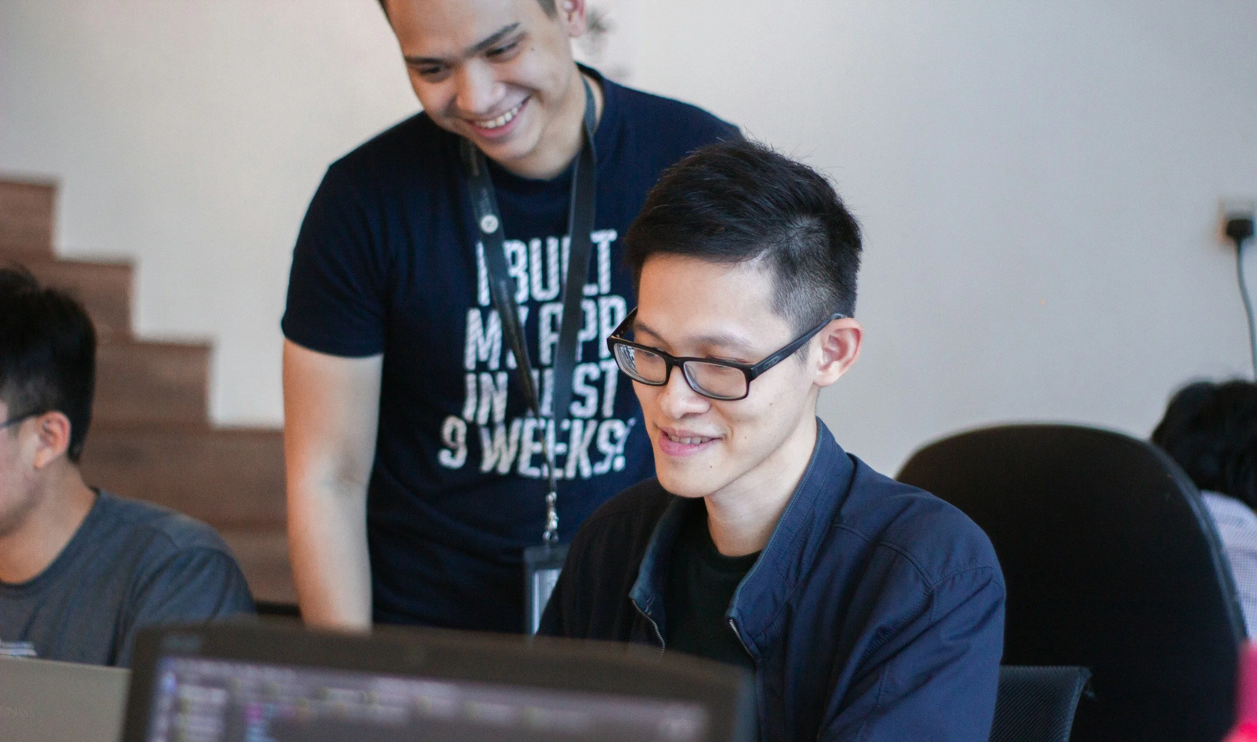 Two people of Asian descent, both with black, short hair. One is wearing glasses and a dark shirt sitting in front of a desk, and the other man is wearing a black shirt and lanyard and standing over the sitting person's right shoulder.