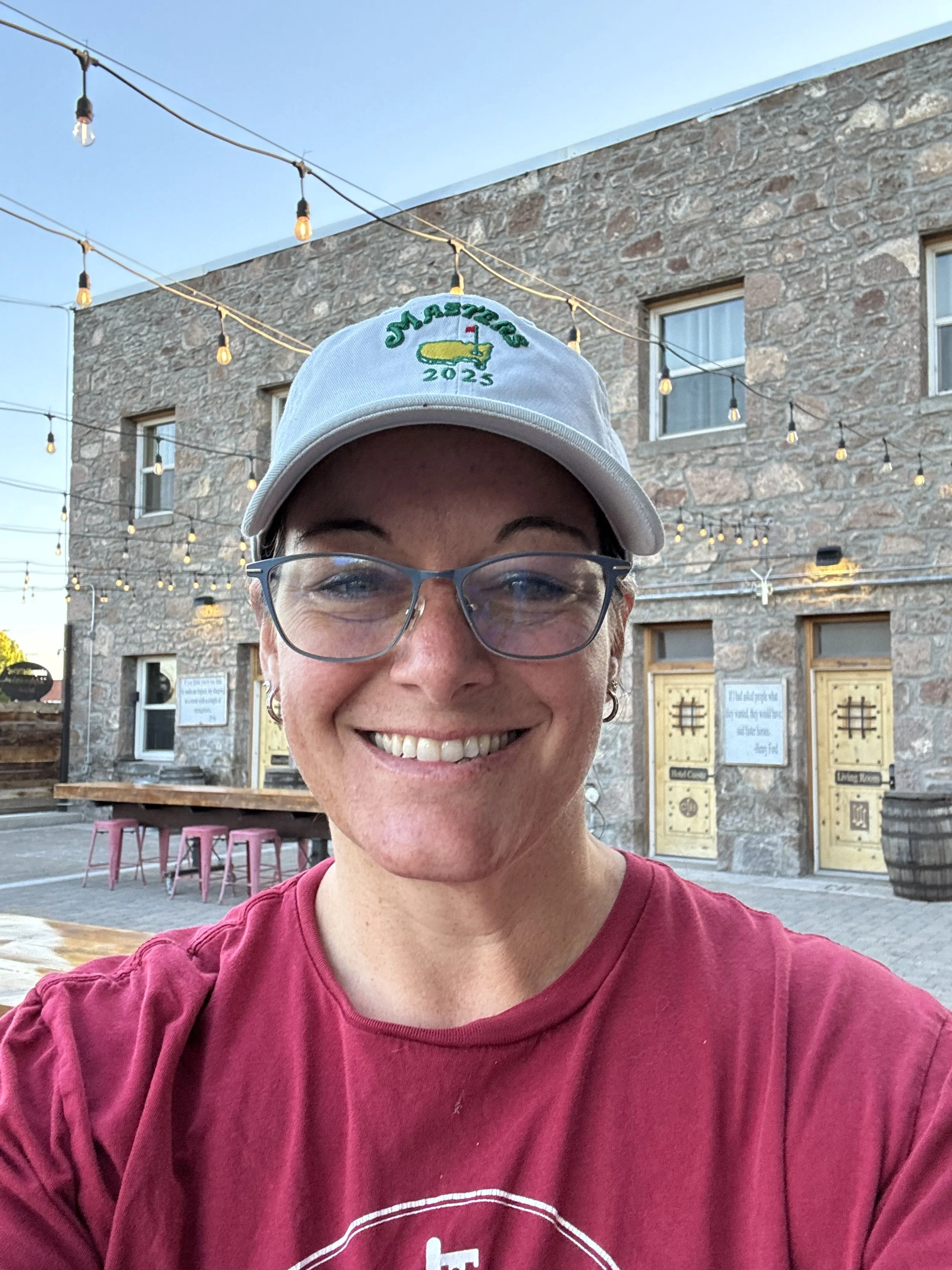 A woman smiling outdoors wearing glasses and a white cap with a daylily flower graphic and the year 2023 on it. Behind her is a rustic stone building with string lights overhead and a wooden bar with pink stools outside.