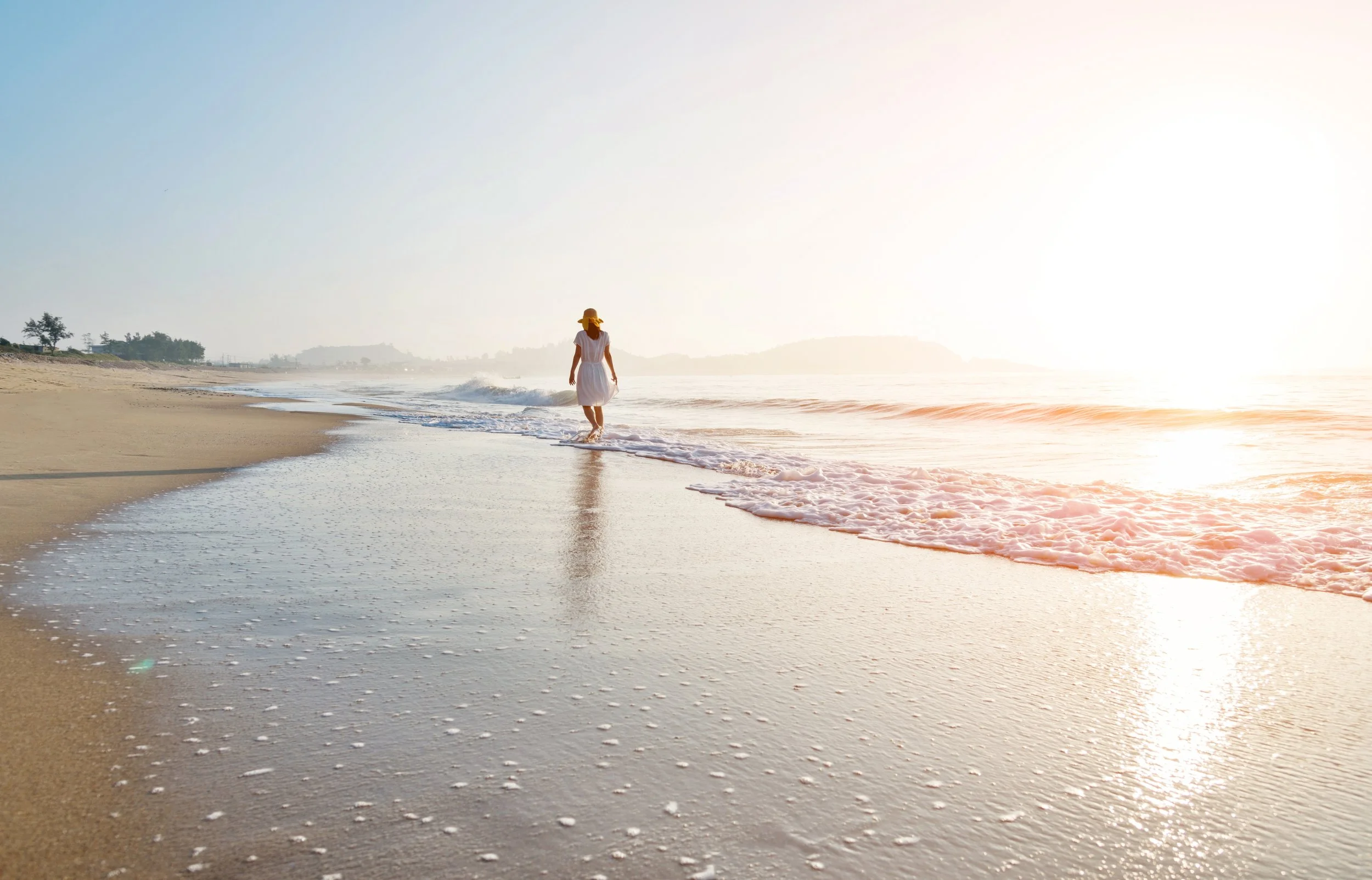 A person walking along the shoreline of a beach at sunset, wearing a white dress and a wide-brimmed hat, with gentle waves washing onto the sand.