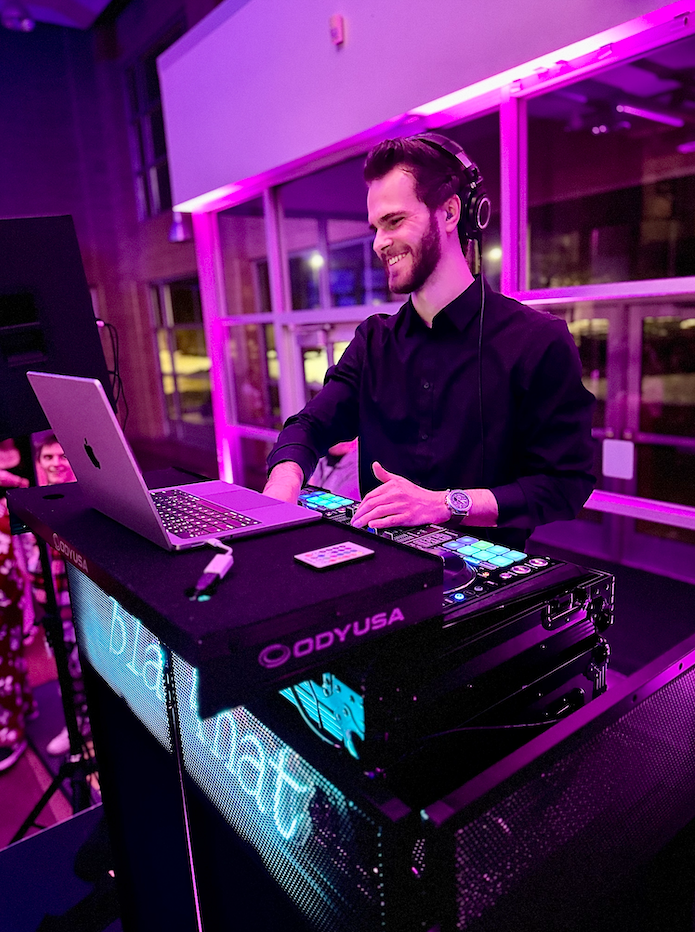 A young man with a beard and headphones, wearing a black shirt, is DJing at a party with purple lighting, using a laptop and DJ equipment.