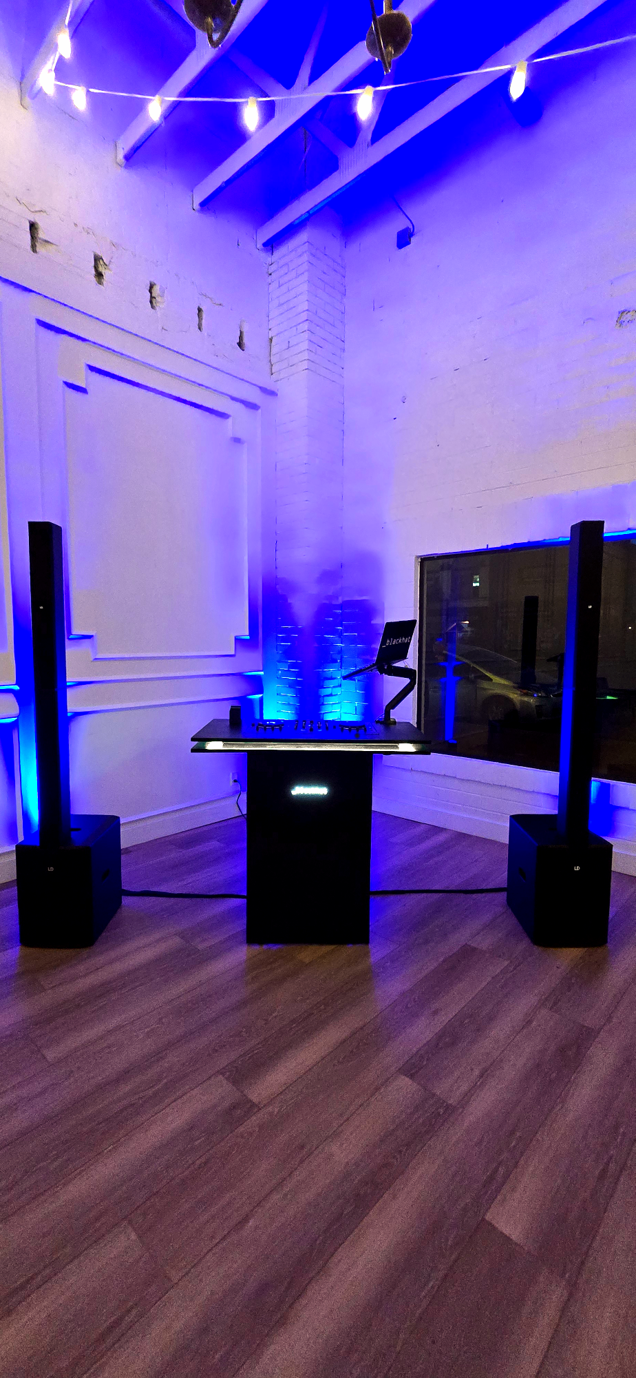 A DJ setup with two large speakers, a mixing console, and a laptop on a small black stand, illuminated by blue LED lighting in a white-walled room with string lights overhead.