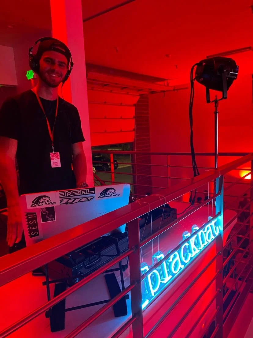 A DJ with headphones standing behind a laptop at a party with a neon sign reading ' blackout' and red lighting.