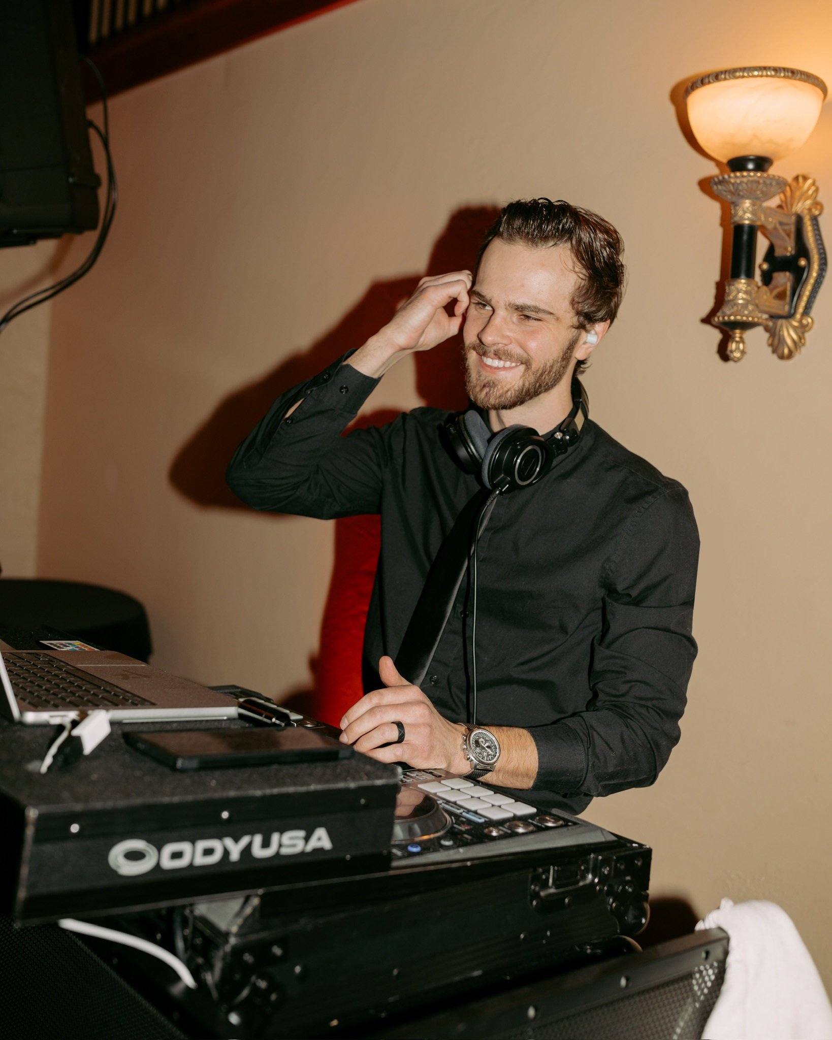 A DJ with a beard, wearing a black shirt, and headphones around his neck, standing at a DJ station with equipment, smiling, and touching his ear, in a warmly lit room.