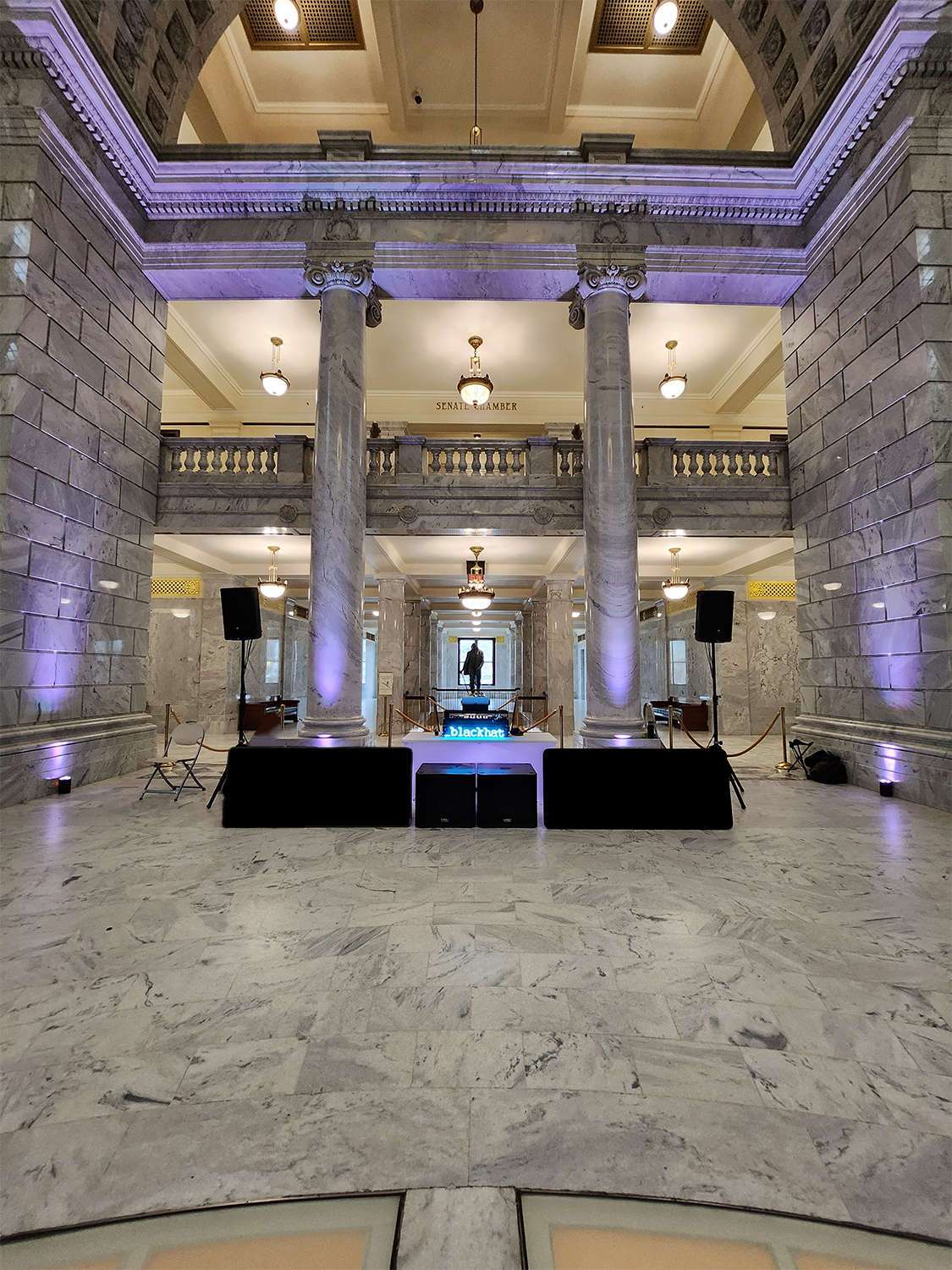 Interior of a grand marble building with columns, balcony, and a small stage with speakers and lighting, set up for a presentation or event.