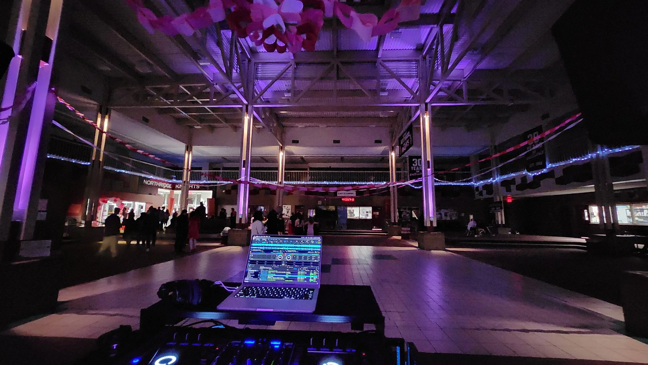 Empty indoor event space or mall with purple and pink lighting, people gathering in the background, DJ equipment with a laptop in the foreground.