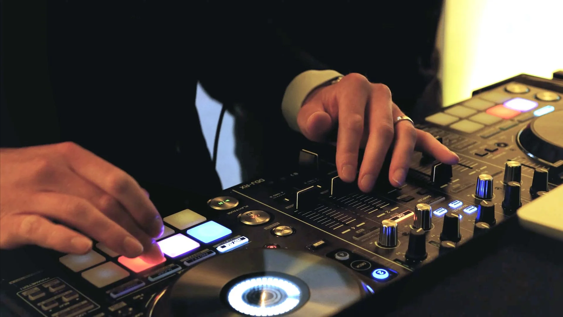 Close-up of a DJ's hands operating a professional DJ mixing console with colorful buttons, knobs, and a turntable.