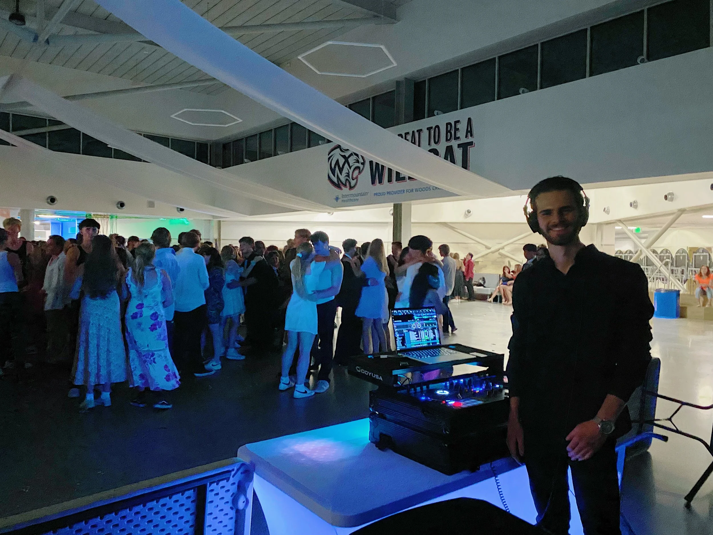 DJ standing next to DJ equipment at an indoor dance party with a large crowd of people dancing in the background.