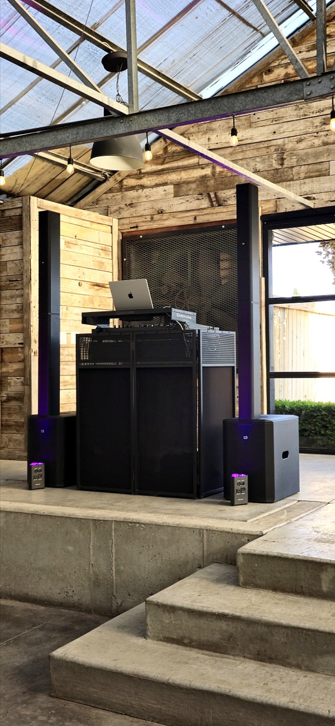 DJ setup with a laptop on a stand, speakers on either side, and colorful string lights in a rustic indoor space with wooden walls and ceiling.