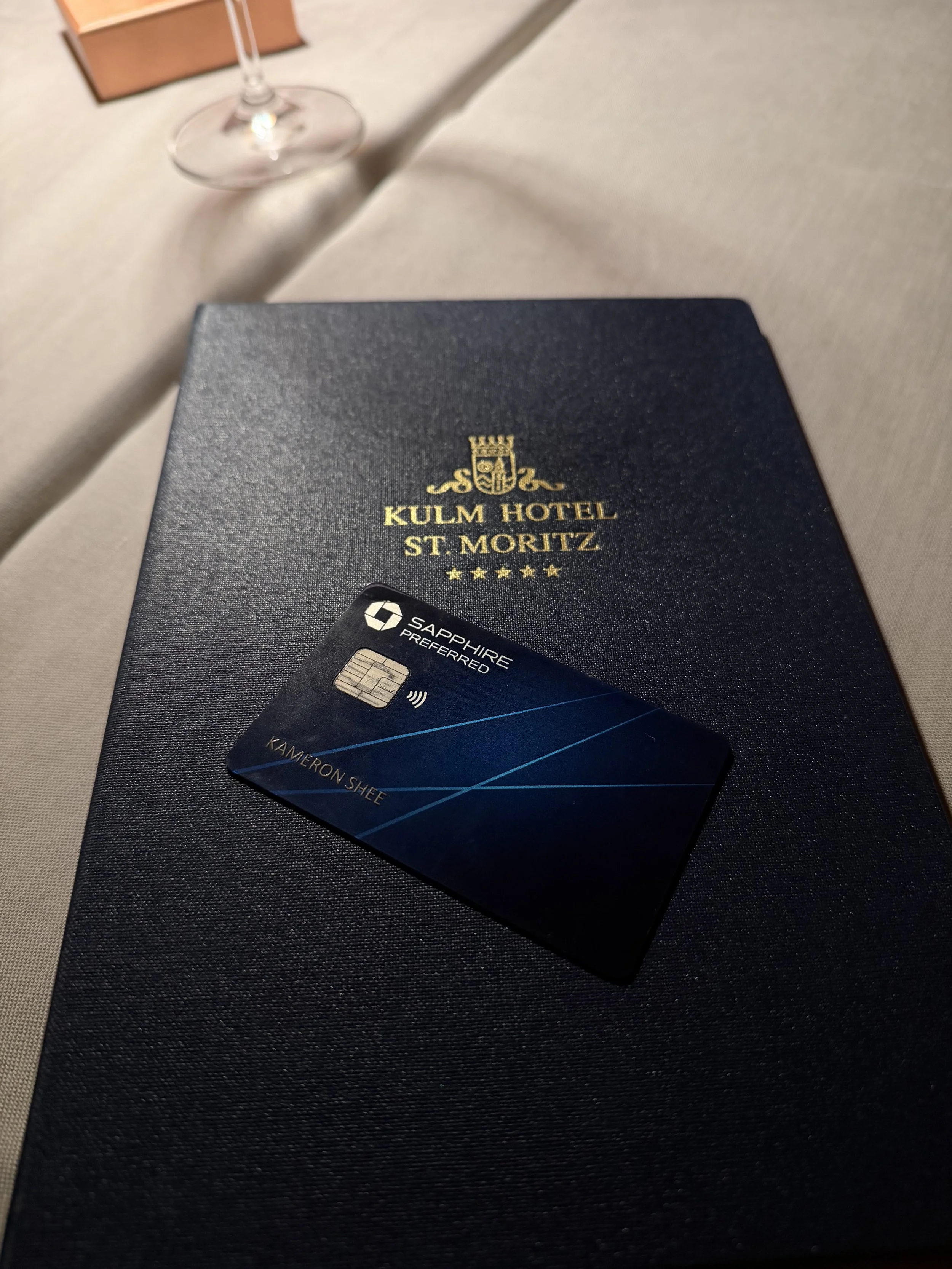 A black card with blue lines and the name 'Kameron Shee' on it, placed on a dark blue booklet labeled 'KULM HOTEL ST. MORITZ' with a crest and four gold stars, on a beige tablecloth with a glass and a wooden box in the background.