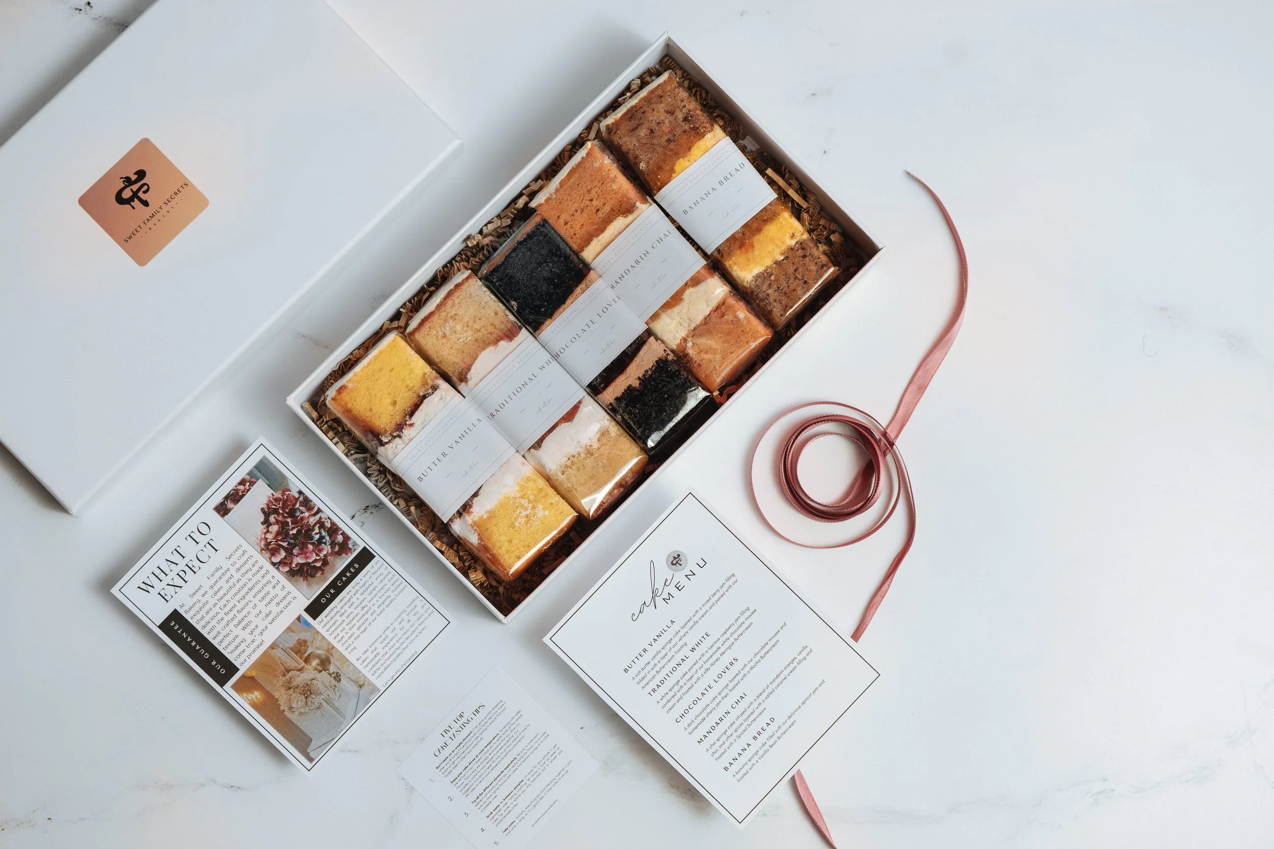 A white box filled with assorted slice cakes labeled with different flavors on a white surface. Next to the box, there is an open menu card and a decorative pink ribbon.