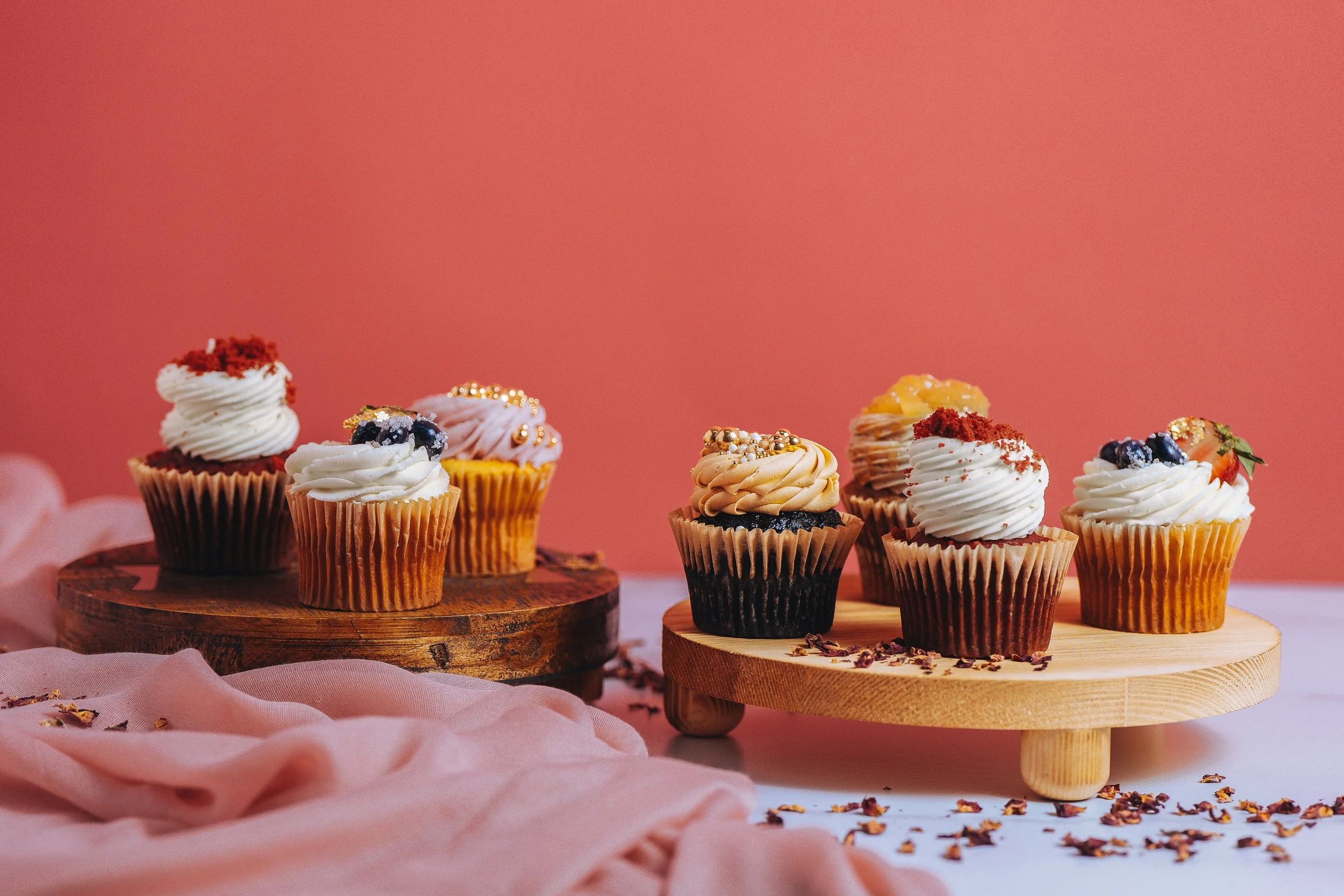 Assorted cupcakes decorated with frosting, berries, and edible florals on wooden stands against a pink background.