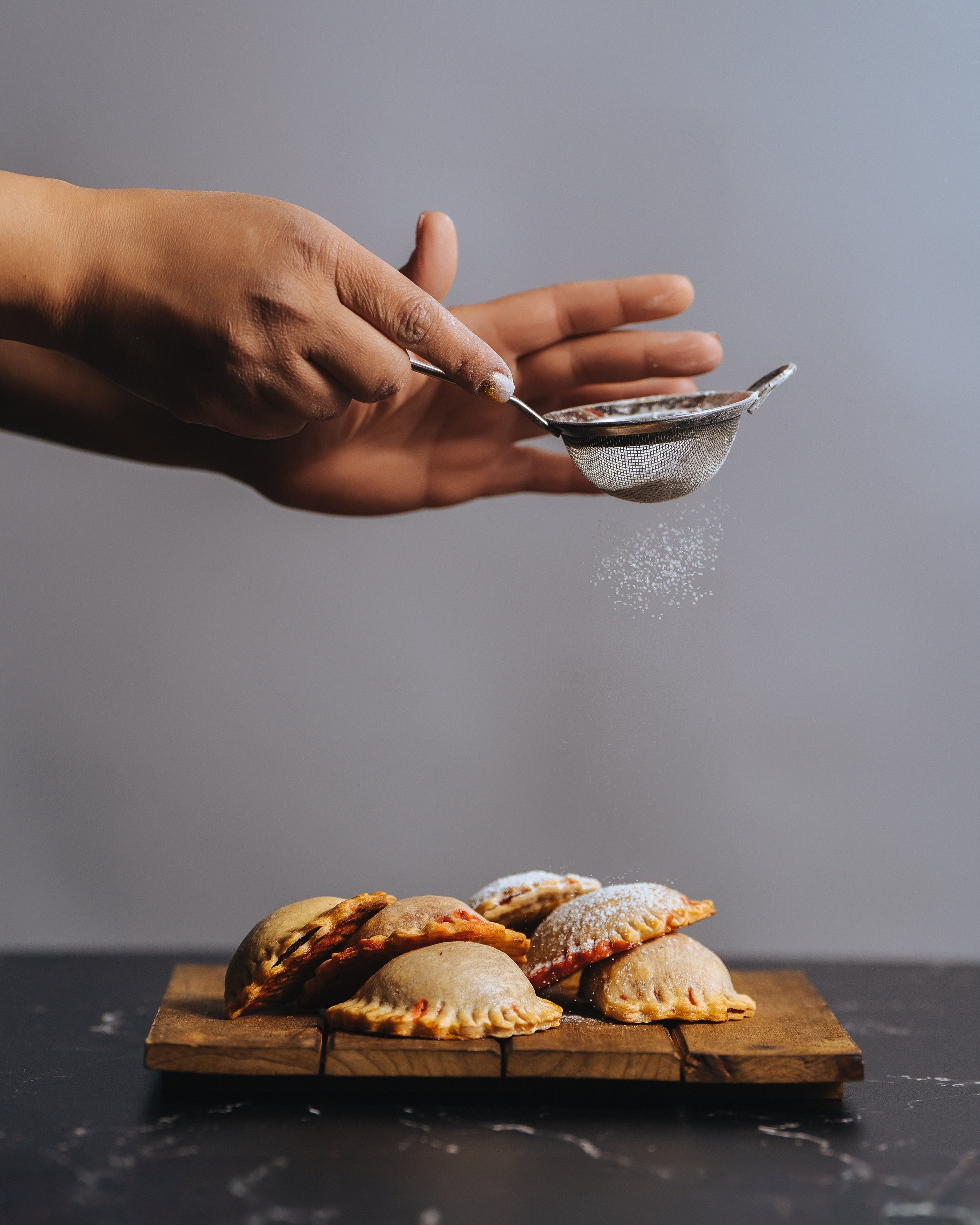 A person dusting powdered sugar onto a plate of baked Jamaican Plantain Tarts on a wooden cutting board.