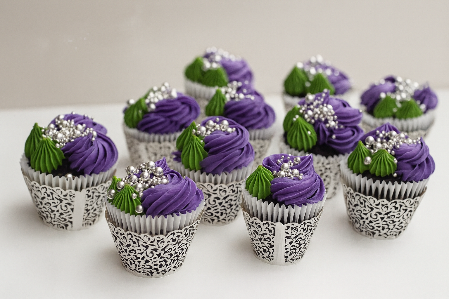 Decorative cupcakes with purple frosting, green leaf-like icing, and silver edible pearls in white lace-patterned wrappers on a white surface.