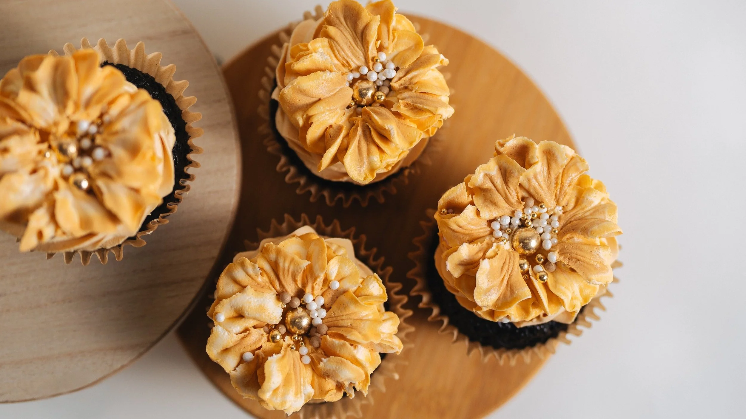 Four cupcakes with piped orange frosting decorated with white, gold, and beige edible pearls on top, placed on a wooden surface.