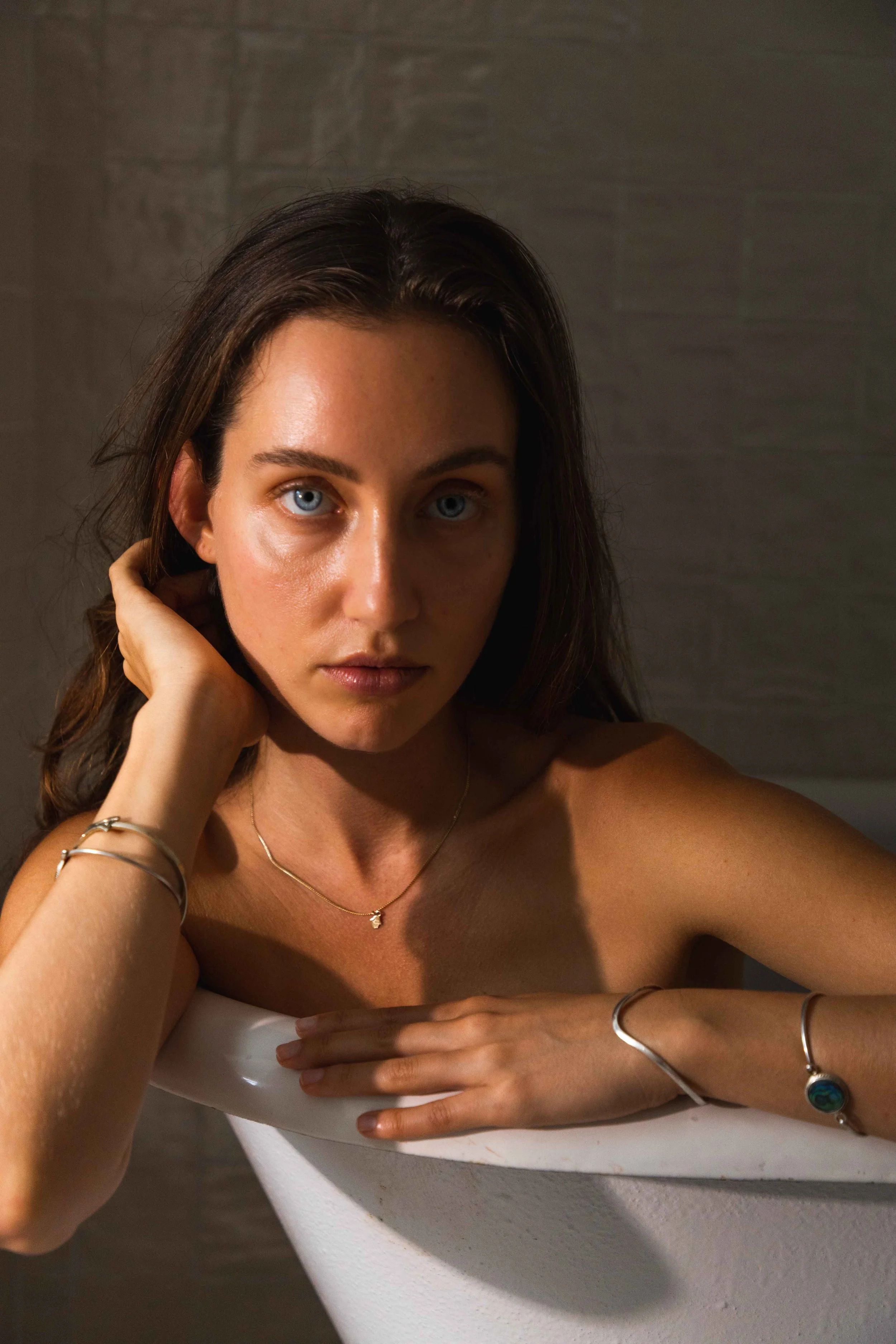 A woman with blue eyes and brown hair resting her arm on the edge of a white bathtub, looking directly at the camera. She is wearing jewelry including bracelets and a necklace, with a neutral background.