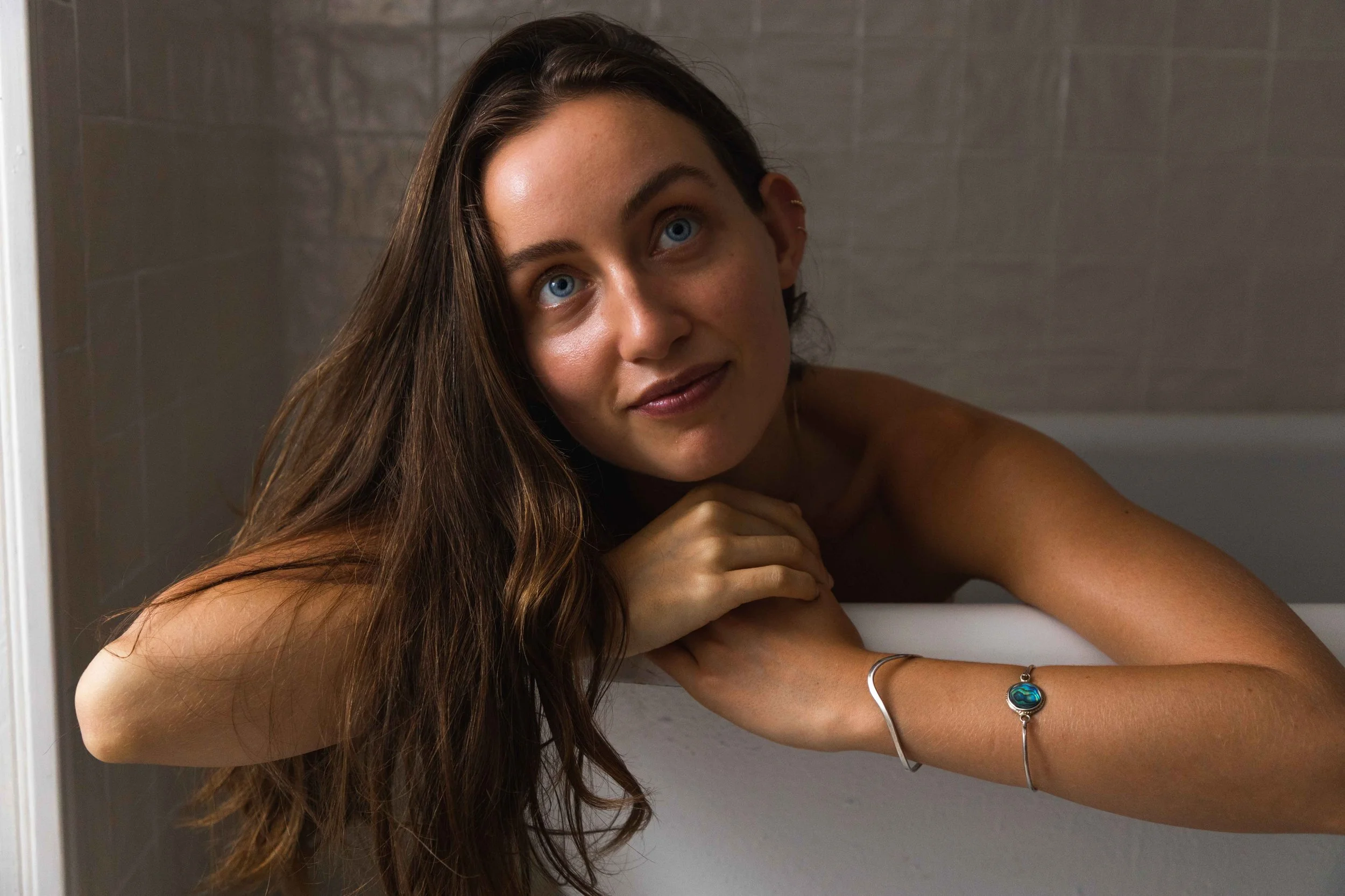 A woman with long brown hair and blue eyes leaning on the edge of a bathtub, looking up at the camera with a gentle smile.