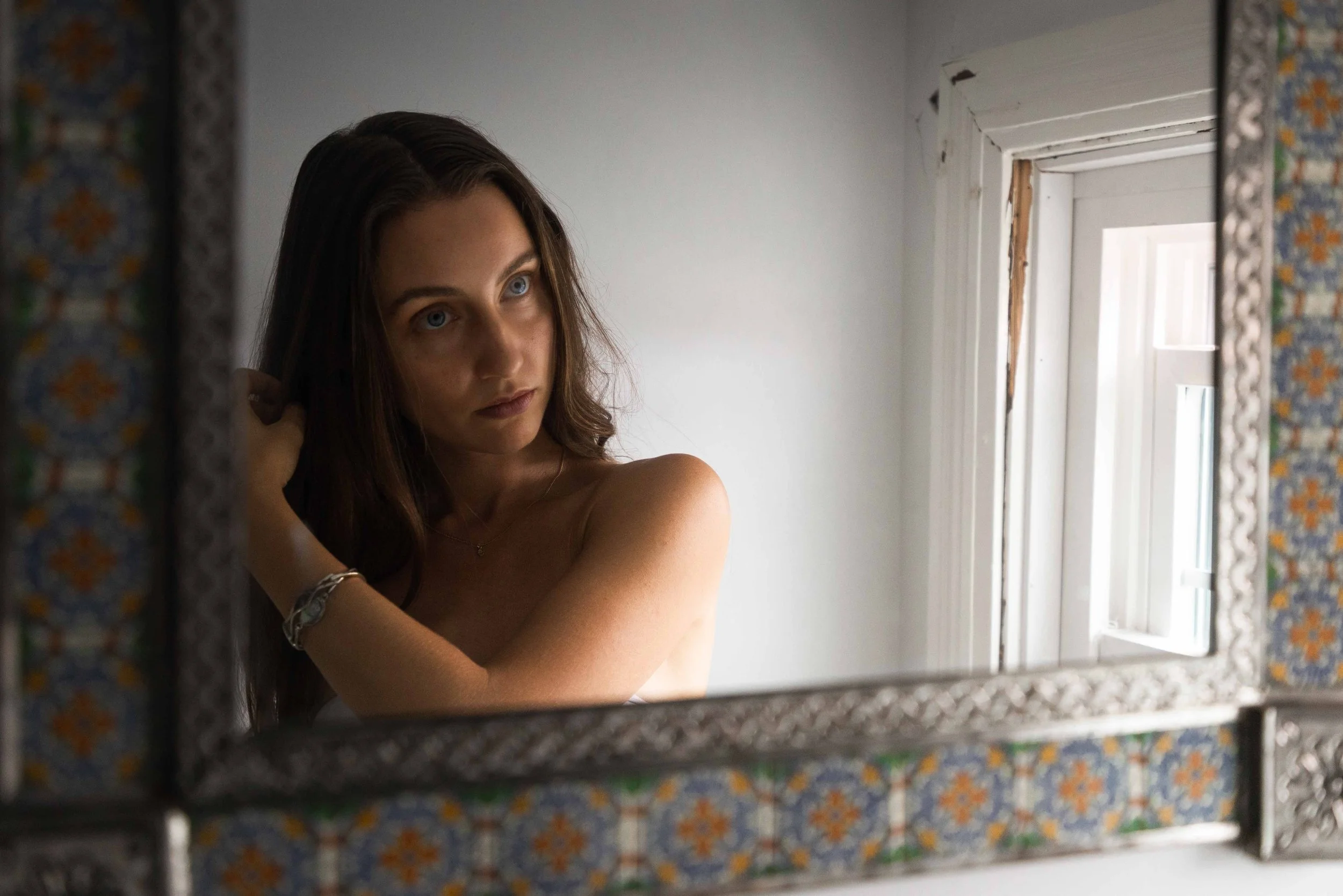 A young woman with long brown hair looking at herself in a mirror, adjusting her hair, with a window reflected in the mirror behind her.