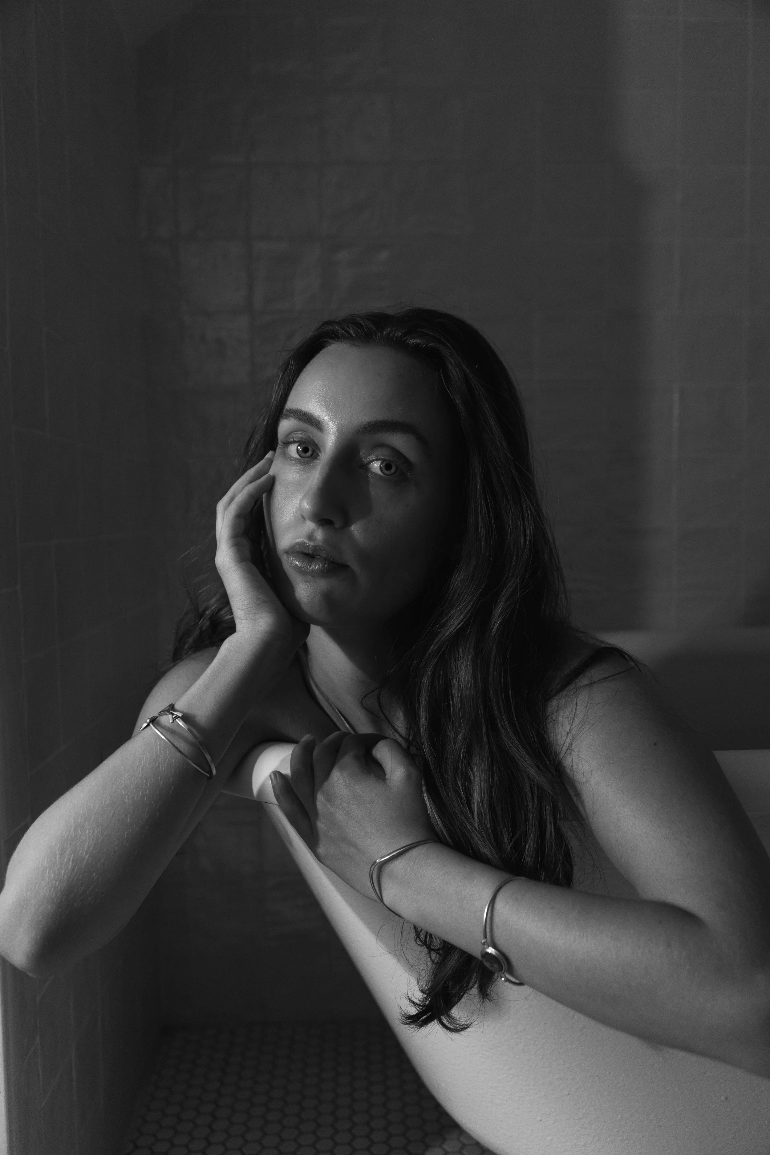 Black and white portrait of a woman with long hair, resting her face on her hand, in a dimly lit room.