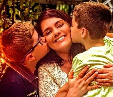 A woman smiling as two children kiss her on the cheeks, one on each side, in an outdoor setting.